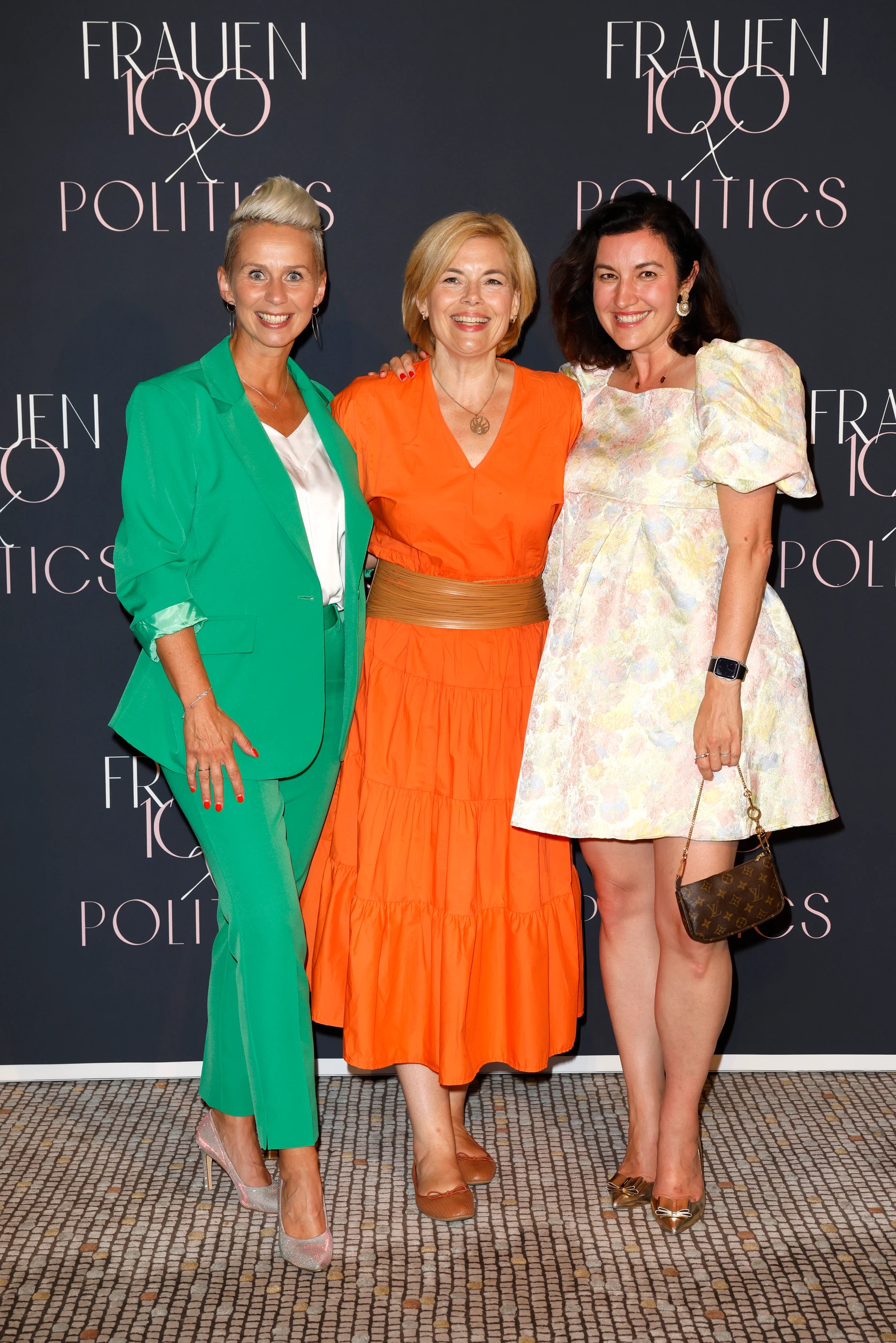 Drei Frauen stehen eng zusammen und lächeln vor einer Fotowand mit der Aufschrift „Frauen100 & Politics“.