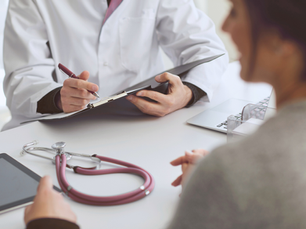 Ärztliche Beratungssituation: Eine Person im weißen Kittel schreibt auf ein Klemmbrett, während eine Patientin gegenüber am Tisch sitzt; auf dem Tisch liegen ein Stethoskop und ein Tablet.