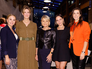 Fünf elegant gekleidete Frauen stehen nebeneinander bei einem Event in einem Innenraum mit warmem Licht und lächeln in die Kamera.