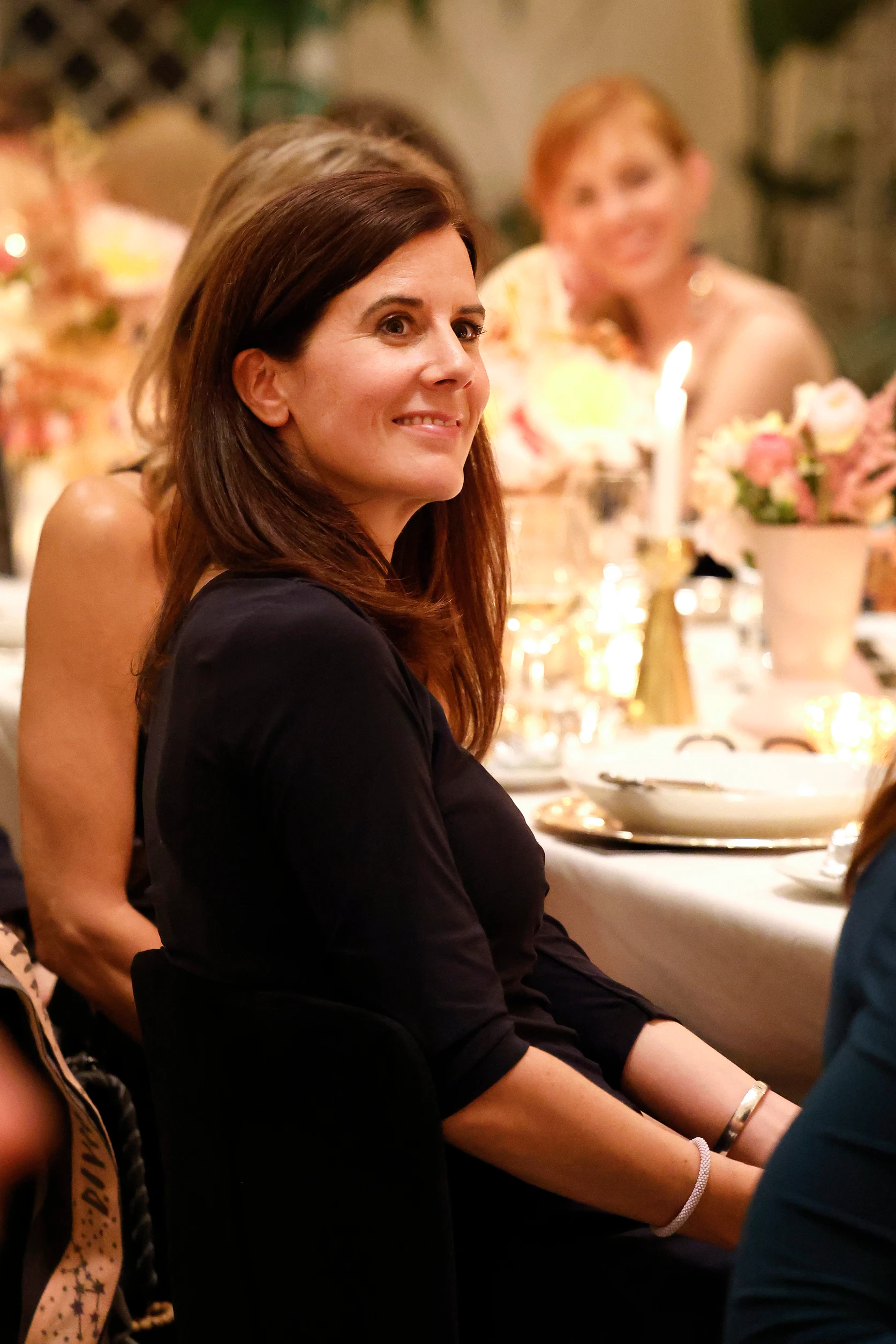 Frau sitzt an einem festlich gedeckten Tisch, dreht sich lächelnd zur Seite, Kerzenlicht und Blumen im Hintergrund.