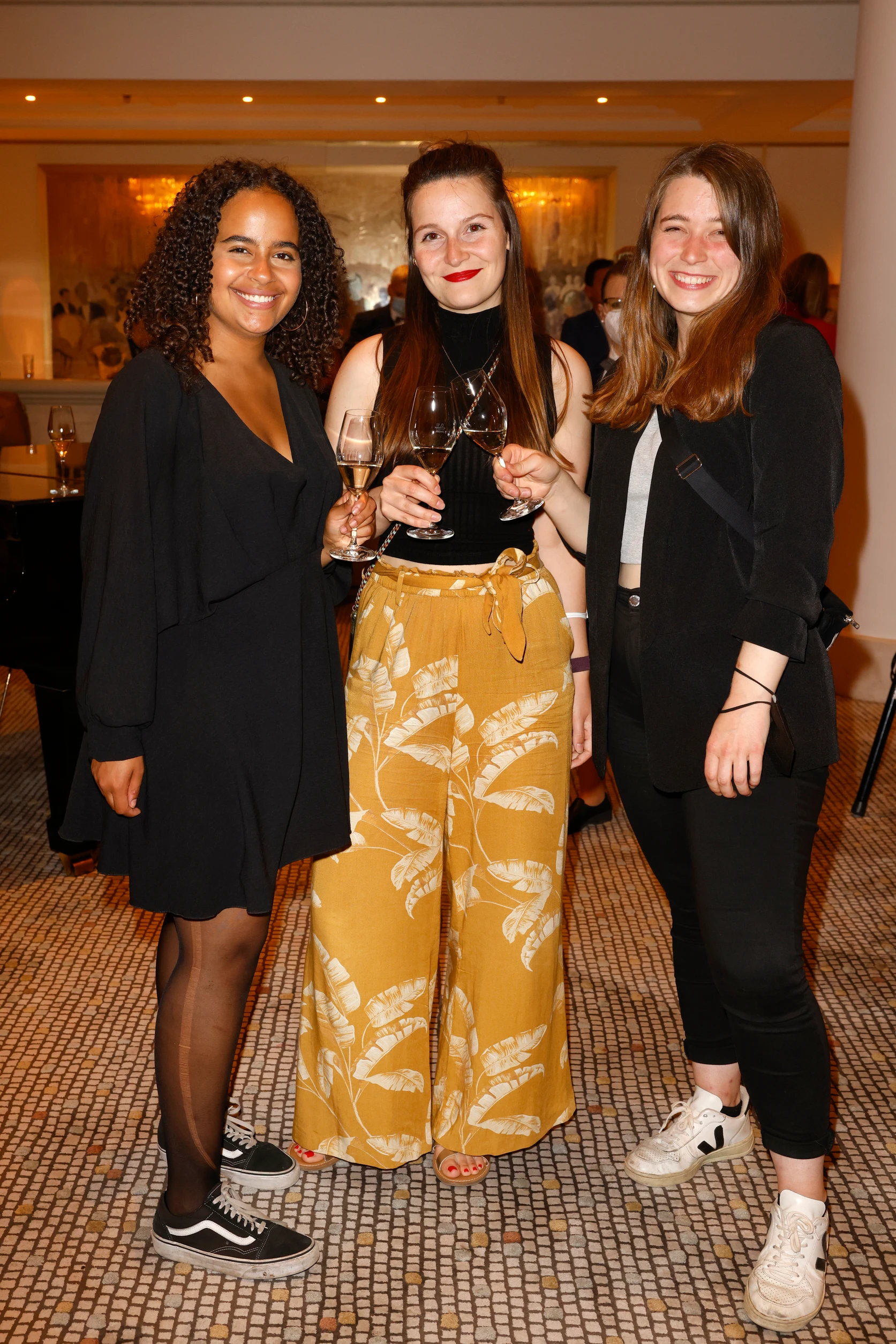 Drei Frauen stehen zusammen, lachen und halten Weingläser bei einem Event.