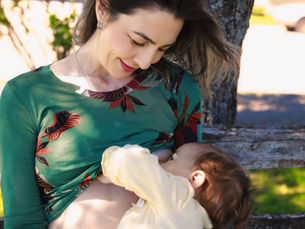 Eine Frau sitzt draußen auf einer Bank und stillt ihr Baby, während sie liebevoll zu ihm hinunterschaut.