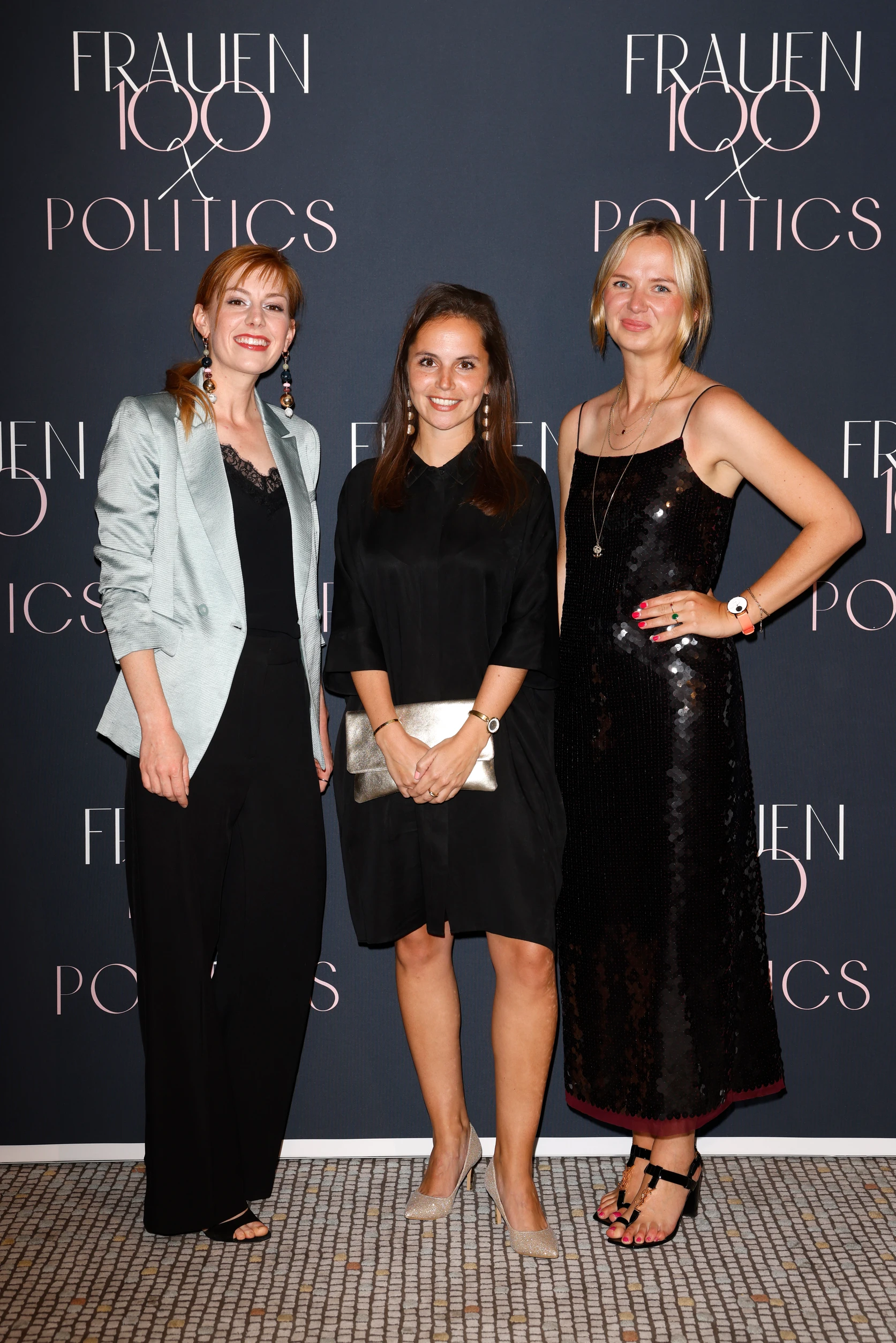 Drei Frauen posieren vor einer dunklen Fotowand mit der Aufschrift „Frauen100 & Politics“.