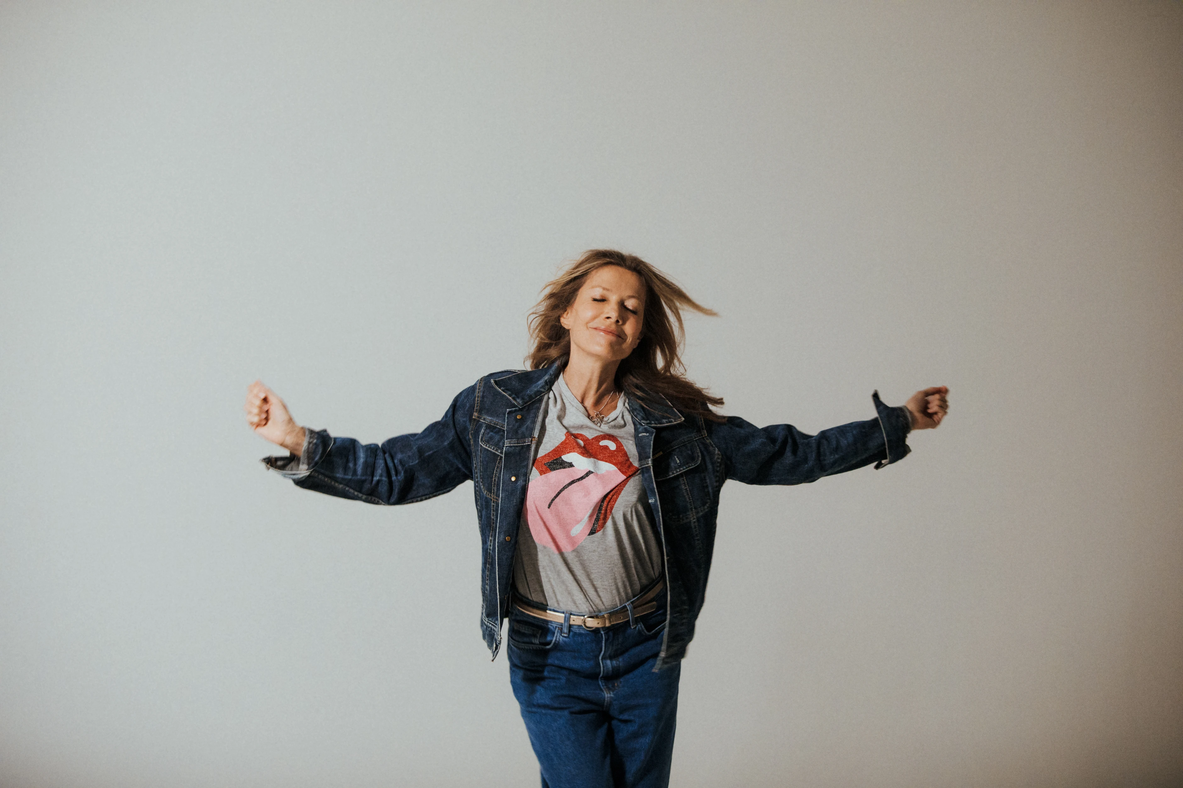Frau im Denim-Outfit mit Rolling-Stones-Shirt bewegt sich dynamisch vor minimalistischem Hintergrund.