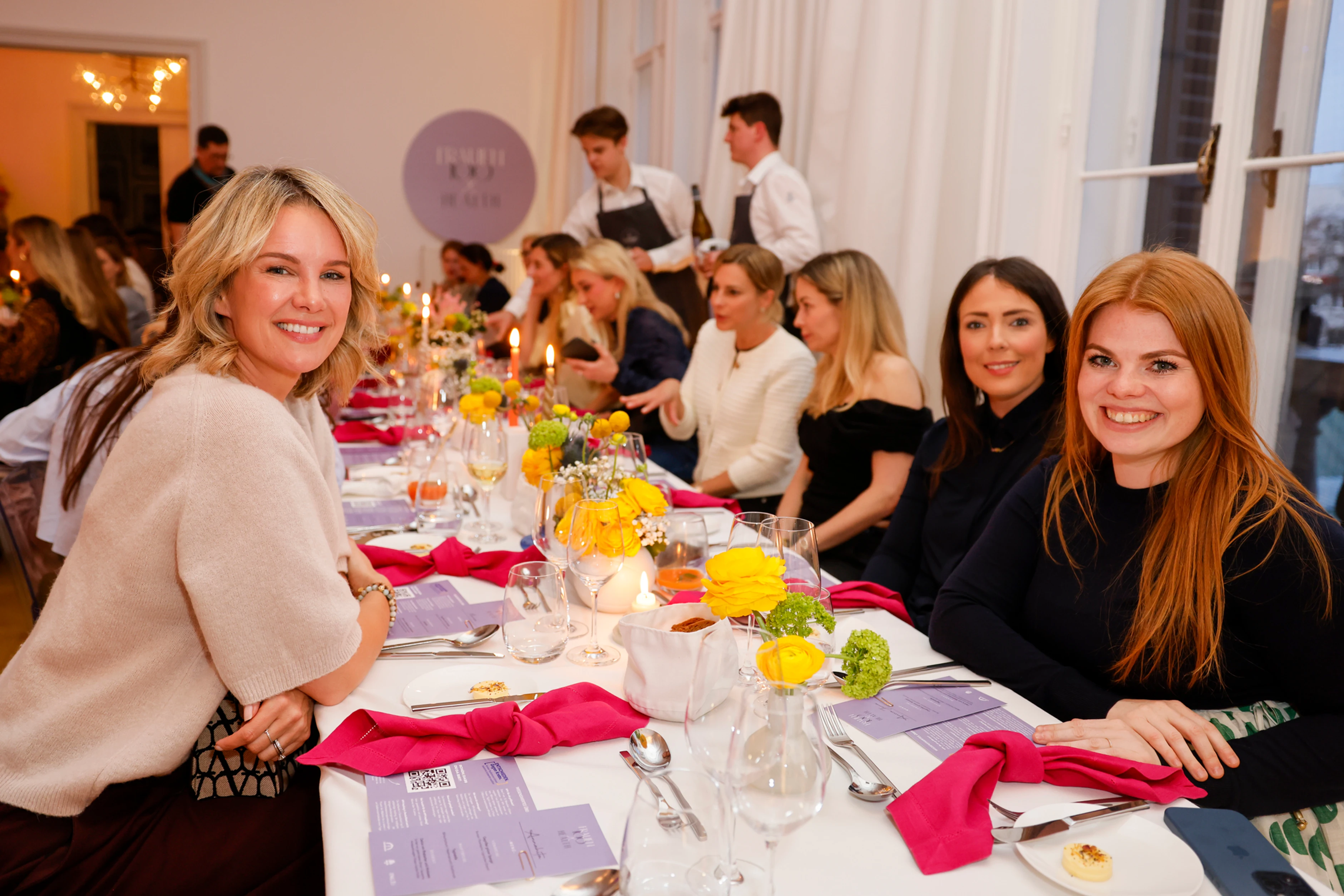 Mehrere Frauen sitzen an einer langen, dekorierten Tafel, einige schauen in die Kamera und lächeln – im Hintergrund serviert das Personal Getränke.