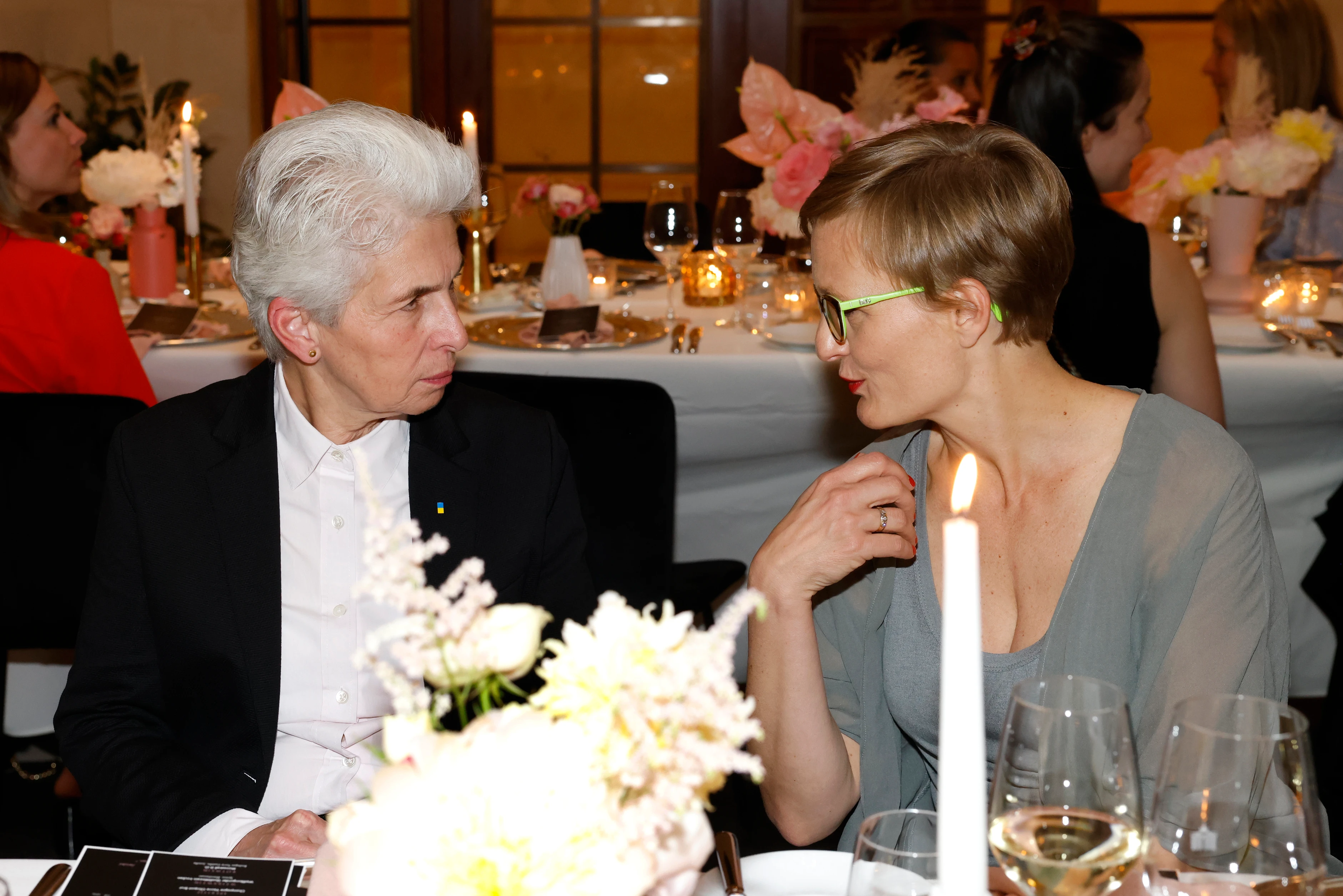 Zwei Frauen sitzen an einem gedeckten Tisch und unterhalten sich, Kerze und Blumen im Vordergrund.