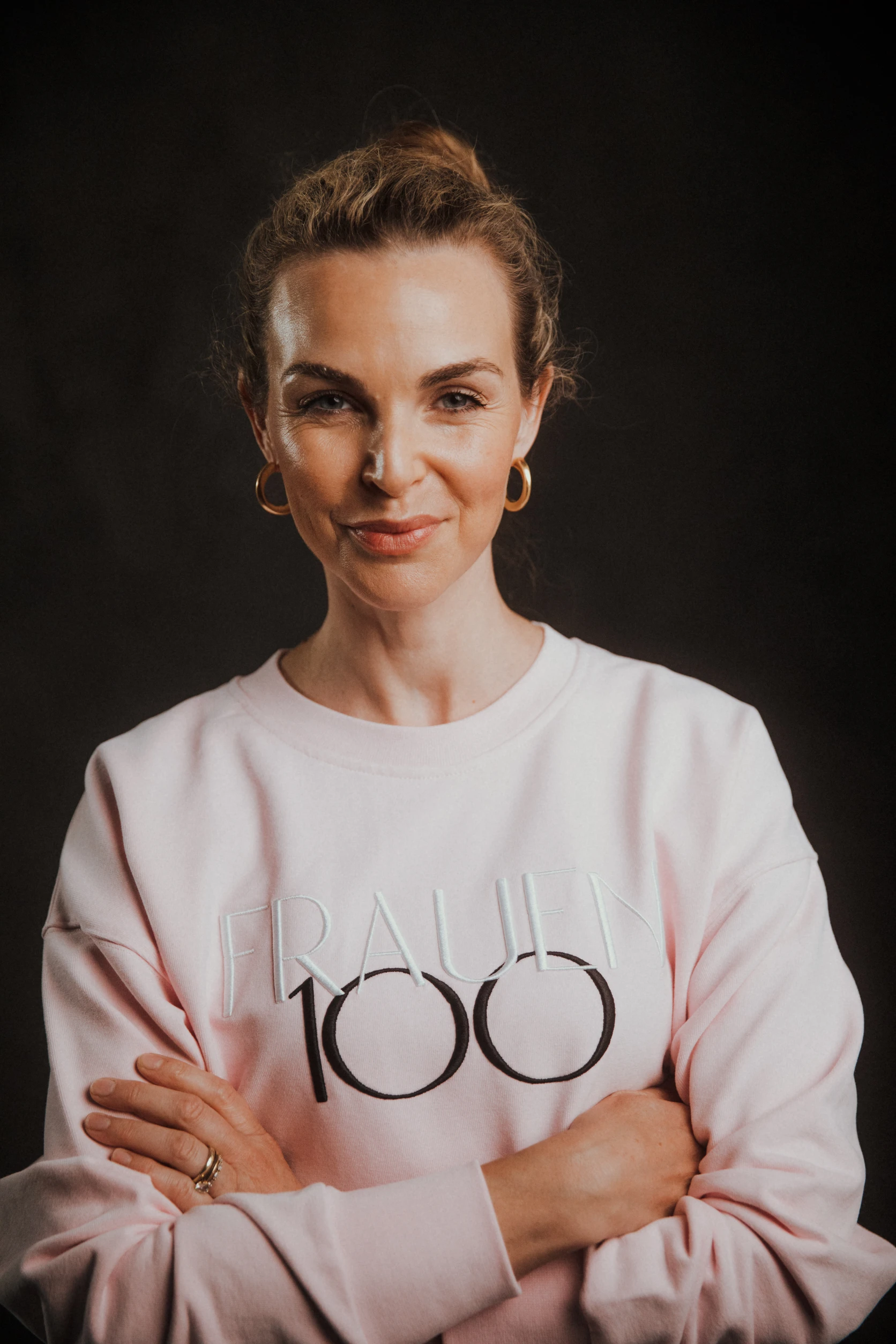 Frau mit zusammengebundenen Haaren trägt rosa „Frauen100“-Sweatshirt und schaut selbstbewusst mit verschränkten Armen in die Kamera.