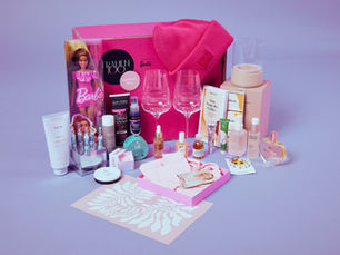 Eine pinke Geschenkbox von FRAUEN100 x Barbie mit verschiedenen Beauty-Produkten, einer Barbie-Puppe, Parfum, Schmuck, zwei Weingläsern und Accessoires ist dekorativ aufgestellt.
