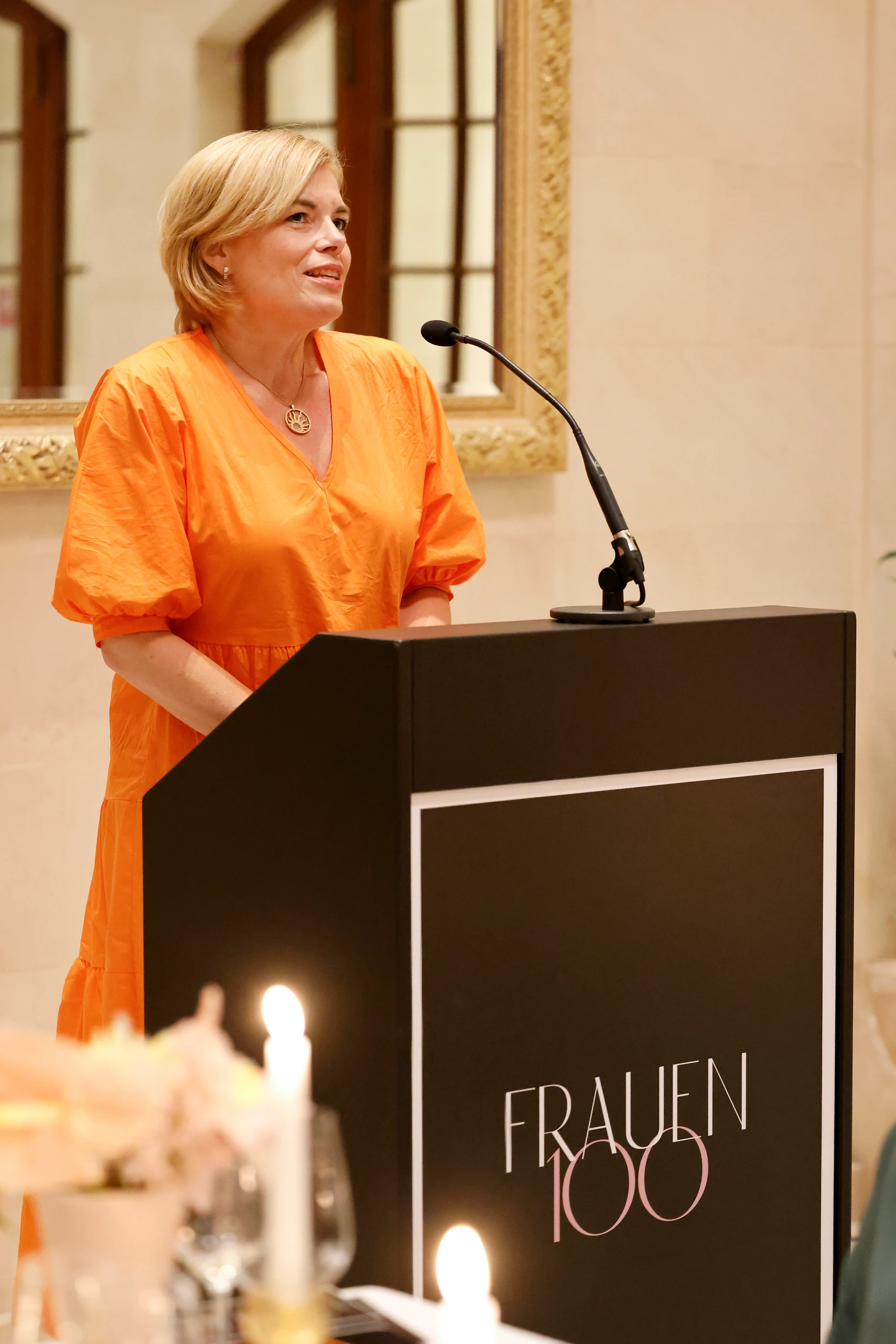 Frau in orangem Kleid spricht an einem Rednerpult bei einer Veranstaltung, Kerzenlicht im Vordergrund.