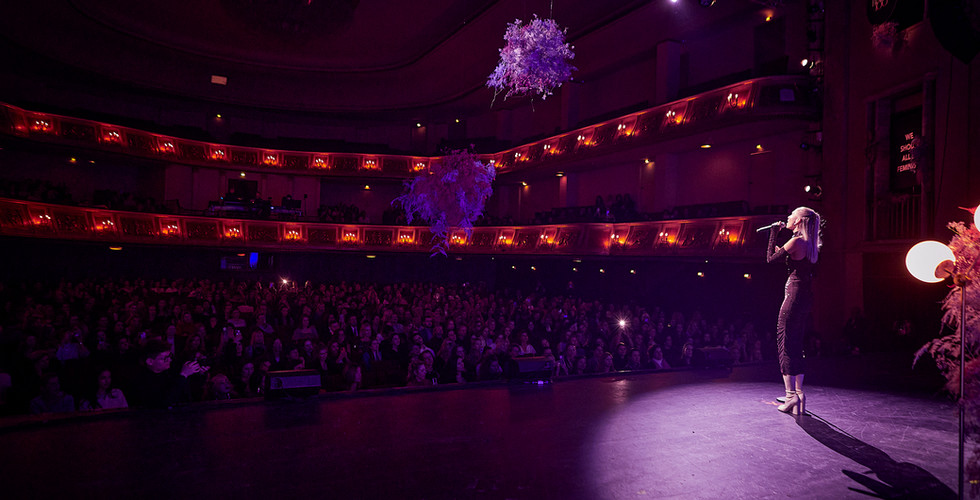 Eine Sängerin steht auf einer Bühne und tritt vor einem großen Publikum in einem Theater mit beleuchteten Rängen auf.