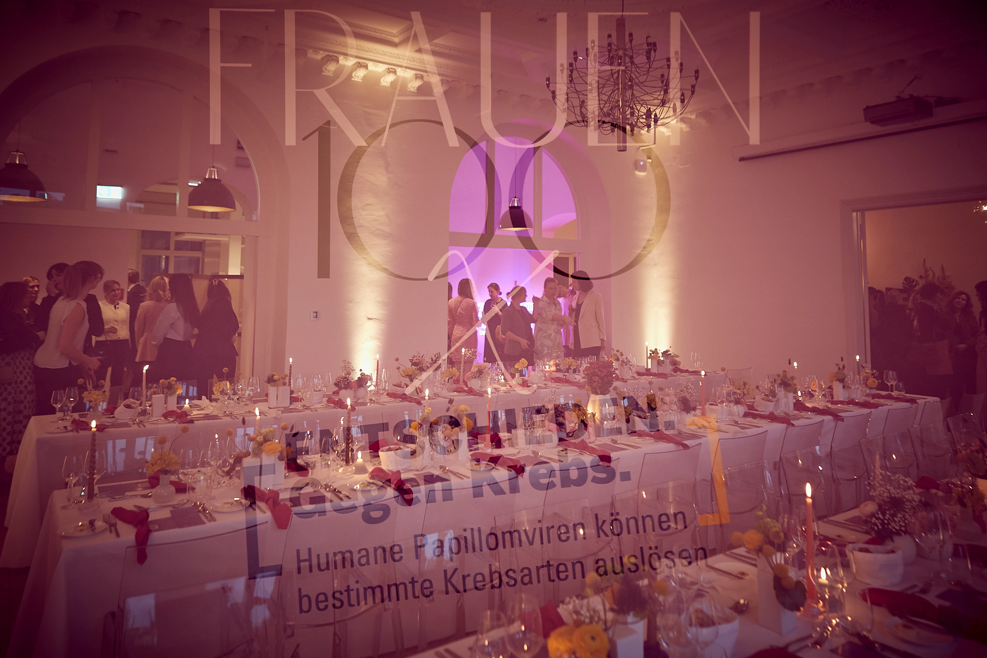 Ein stilvoll gedeckter Saal mit langen Tafeln, Kerzenlicht und Blumenarrangements – Gäste bewegen sich im Raum, während im Hintergrund die „Frauen 100“-Botschaft durchscheint.