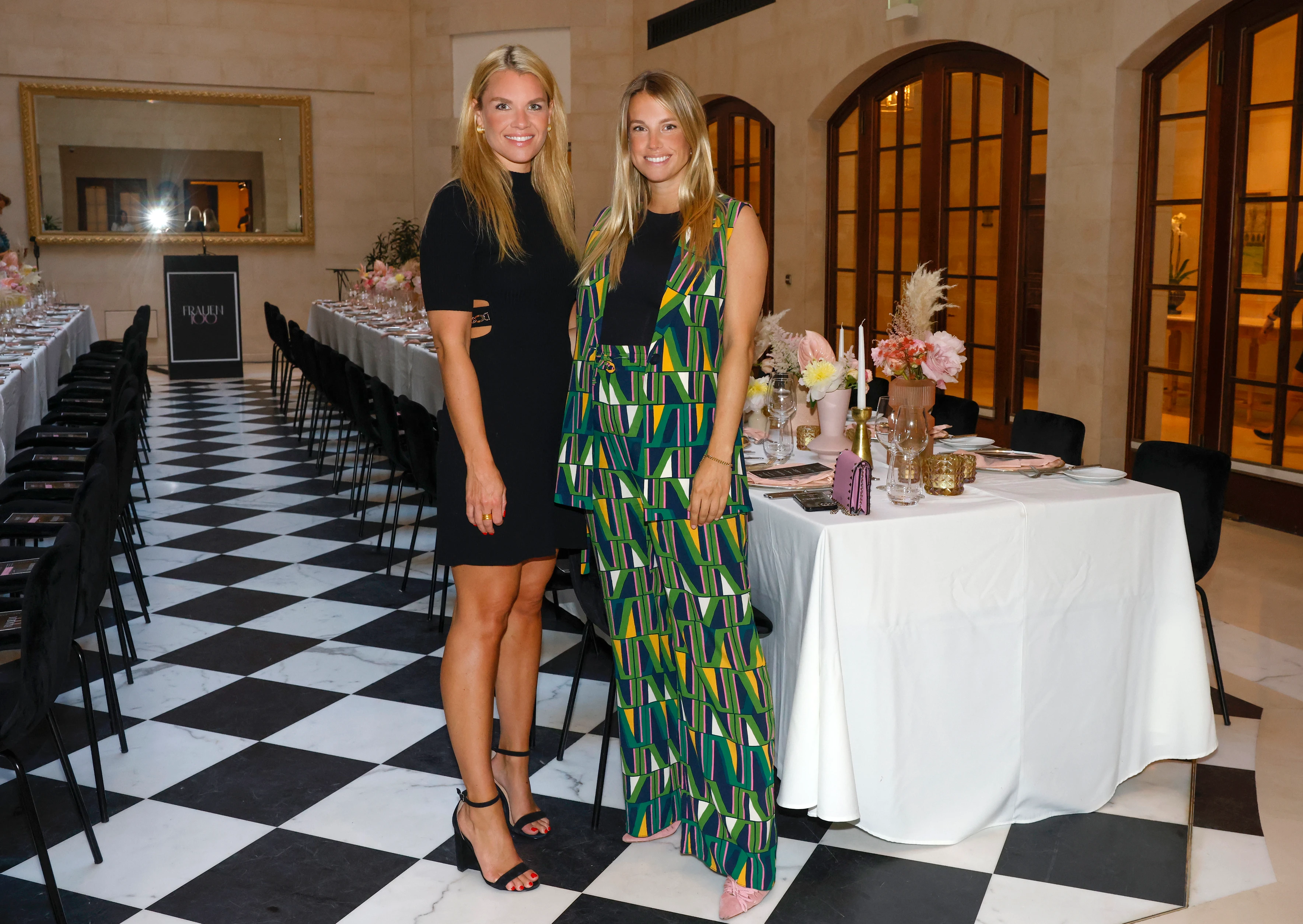 Zwei Frauen posieren in einem festlich dekorierten Saal mit langen gedeckten Tischen und Blumenarrangements.