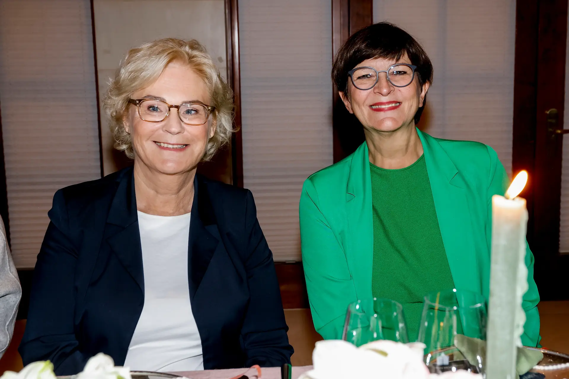 Zwei Frauen sitzen an einem Tisch, lächeln und schauen in die Kamera, vor ihnen stehen Gläser und eine Kerze.