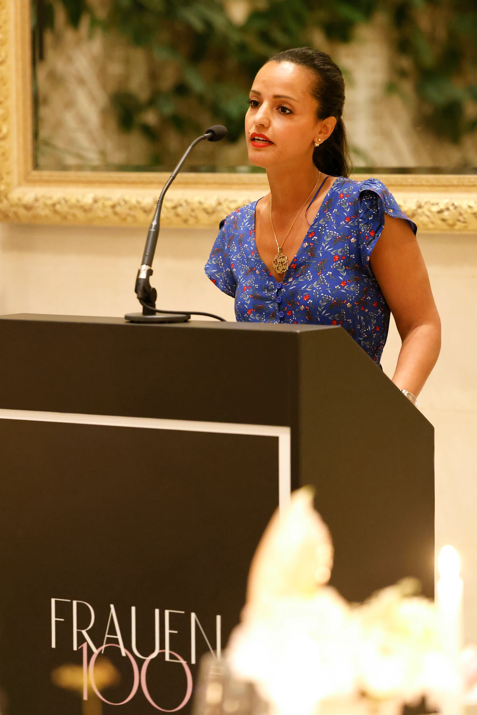 Frau in blauem Kleid spricht an einem Rednerpult mit Mikrofon bei einer Veranstaltung „Frauen100“.