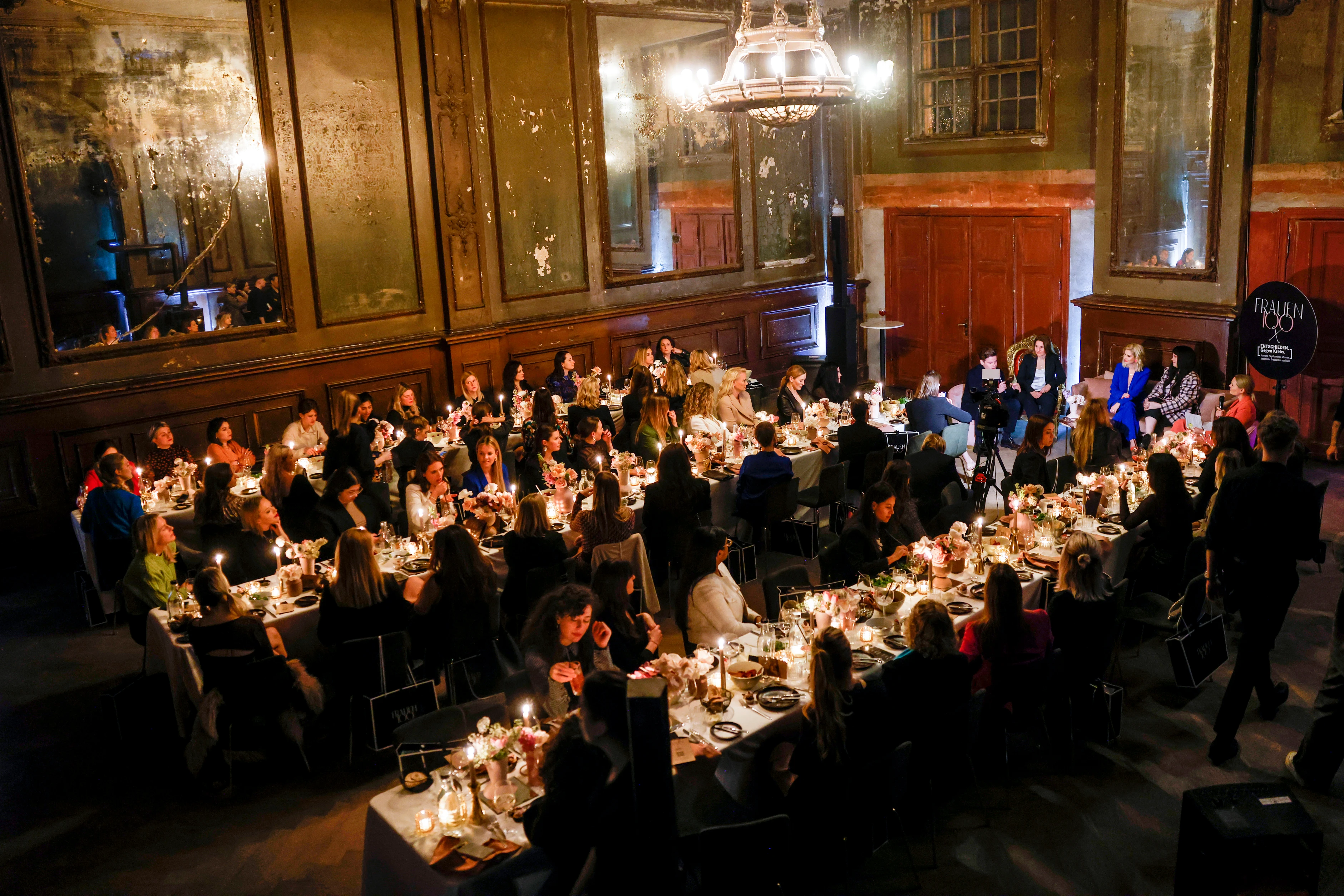 Eine große Eventlocation mit gedeckten Tischen und vielen Gästen während einer Paneldiskussion.