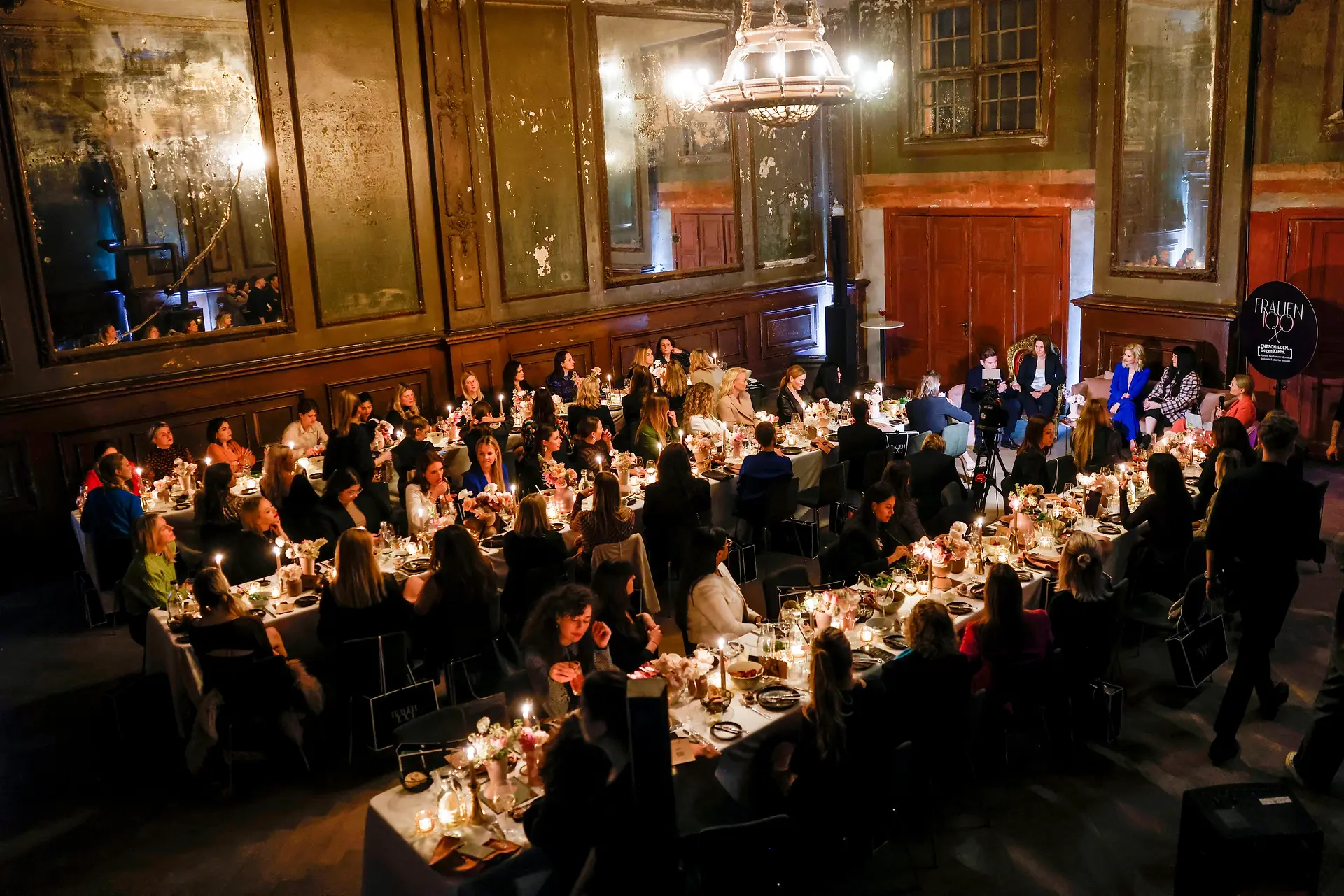 Eine große Eventlocation mit gedeckten Tischen und vielen Gästen während einer Paneldiskussion.