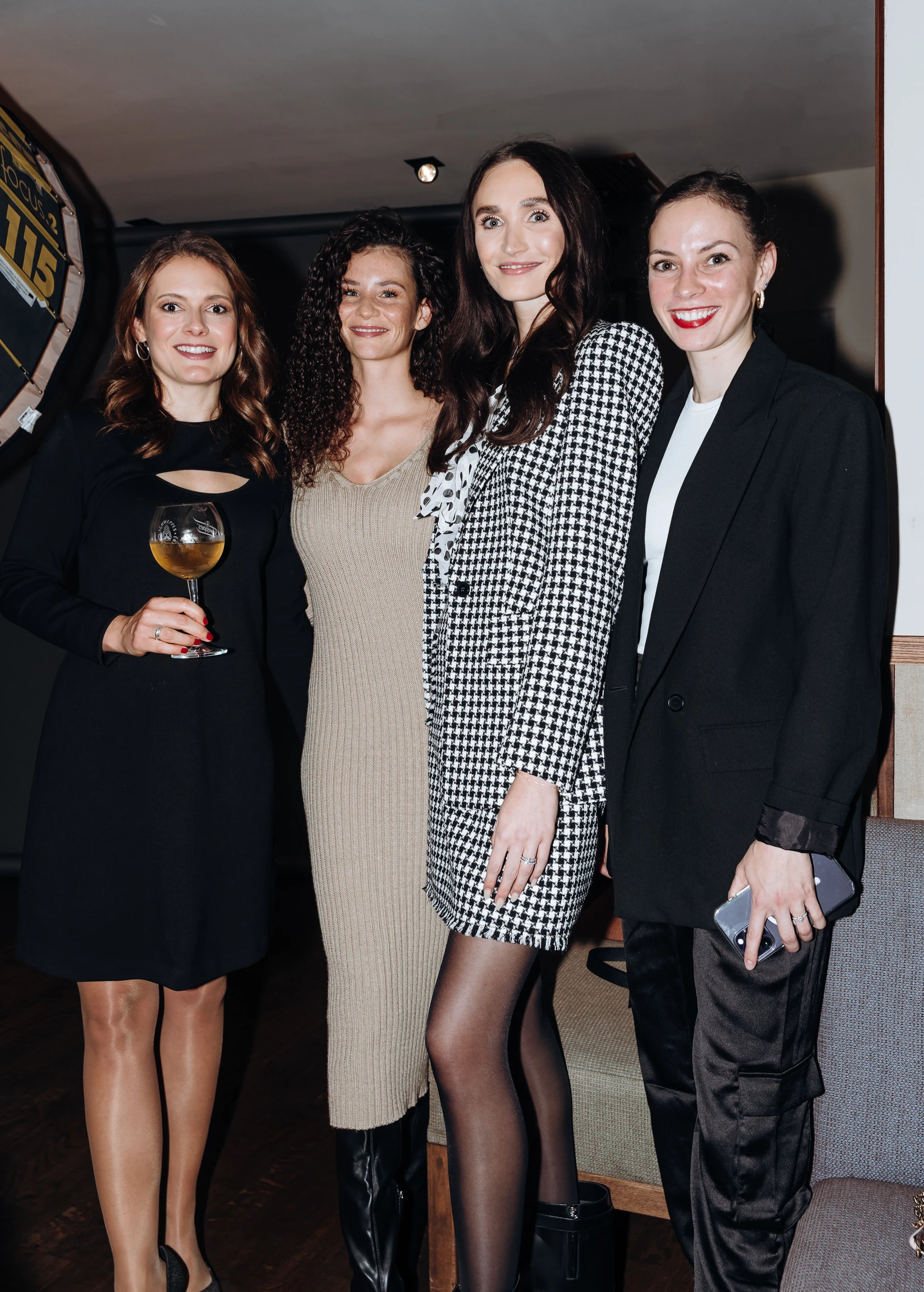 Vier Frauen posieren gemeinsam bei einem Event, eine hält ein Weinglas, alle tragen elegante Abendkleidung.