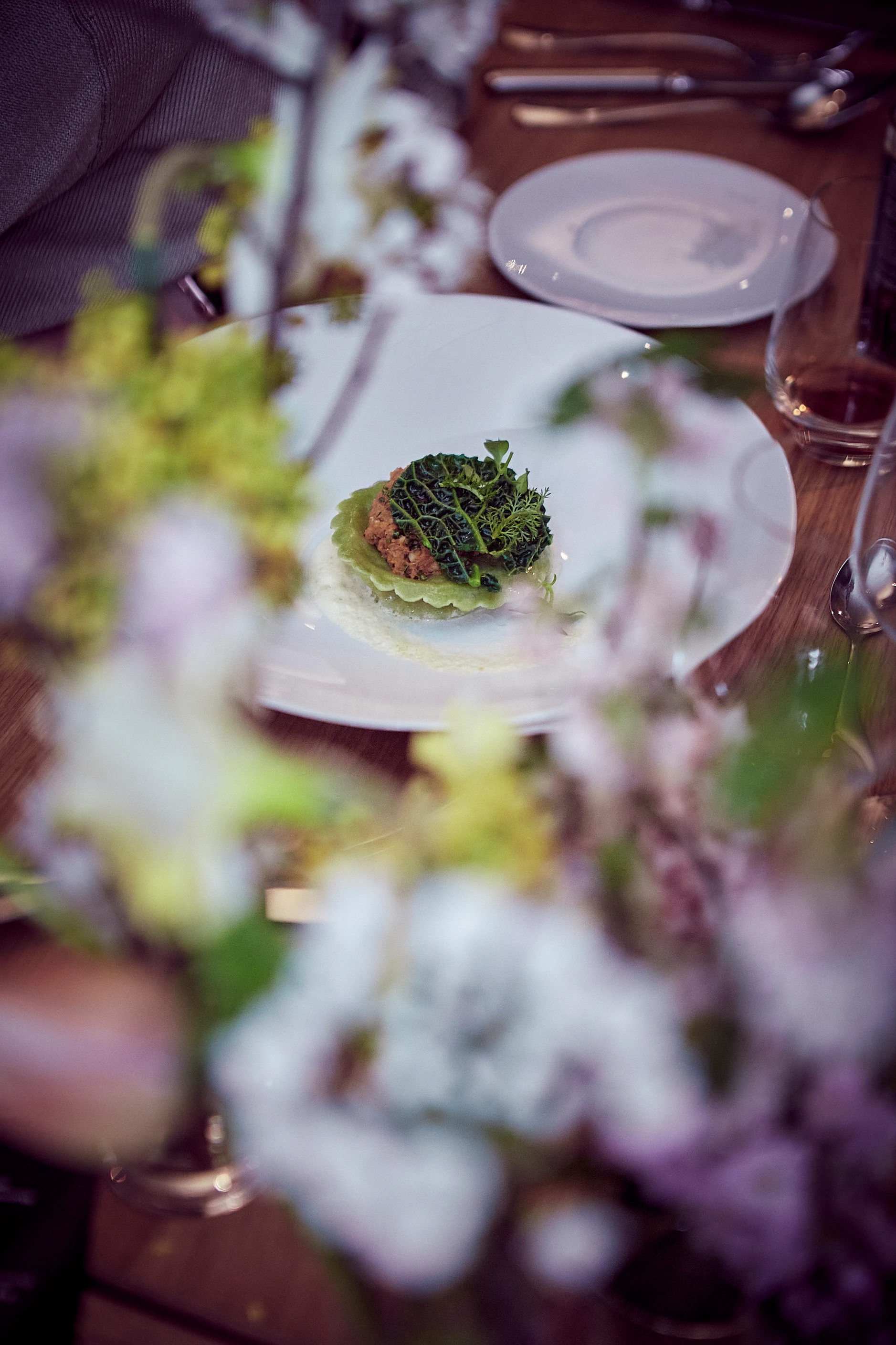 Ein angerichteter Teller mit einer kleinen Speise steht auf einem gedeckten Tisch, umgeben von Geschirr und Blumen.