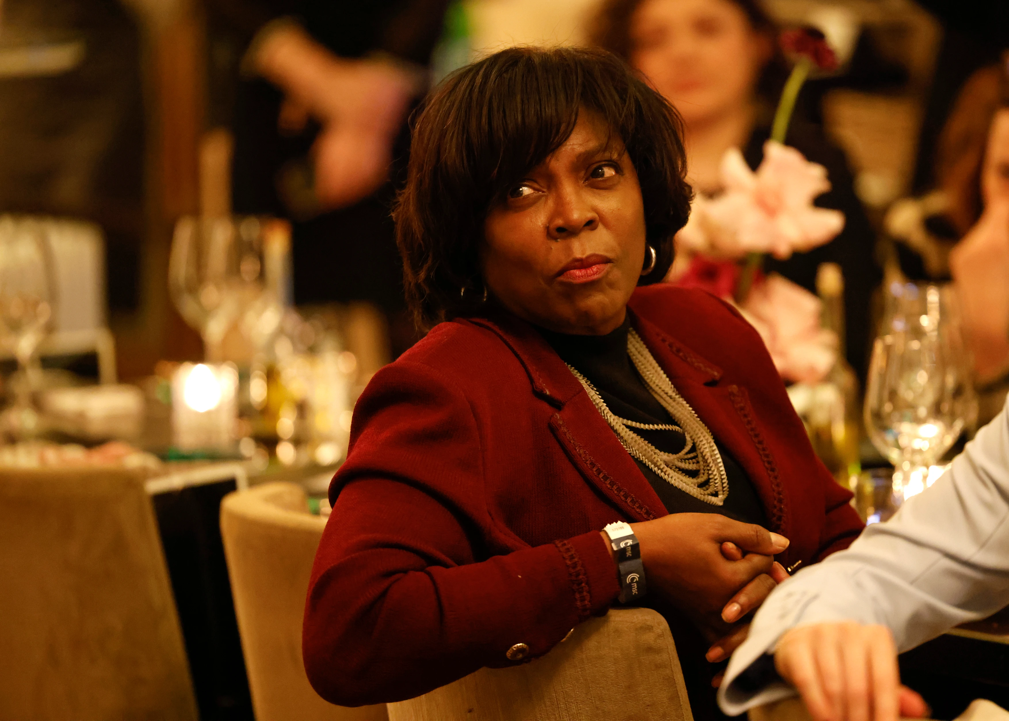 Frau in rotem Blazer sitzt bei einem Event an einem gedeckten Tisch und blickt aufmerksam zur Seite, im Hintergrund warmes Kerzenlicht und Gäste. 