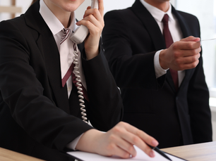 Zwei Personen in Businesskleidung an einem Schreibtisch: Eine Person telefoniert mit einem Festnetztelefon, während die andere eine Karte oder ein Dokument überreicht.