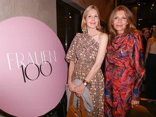 Zwei Frauen posieren lächelnd bei einem Event vor einem großen runden Schild mit der Aufschrift „FRAUEN 100“, sie tragen elegante gemusterte Kleider.