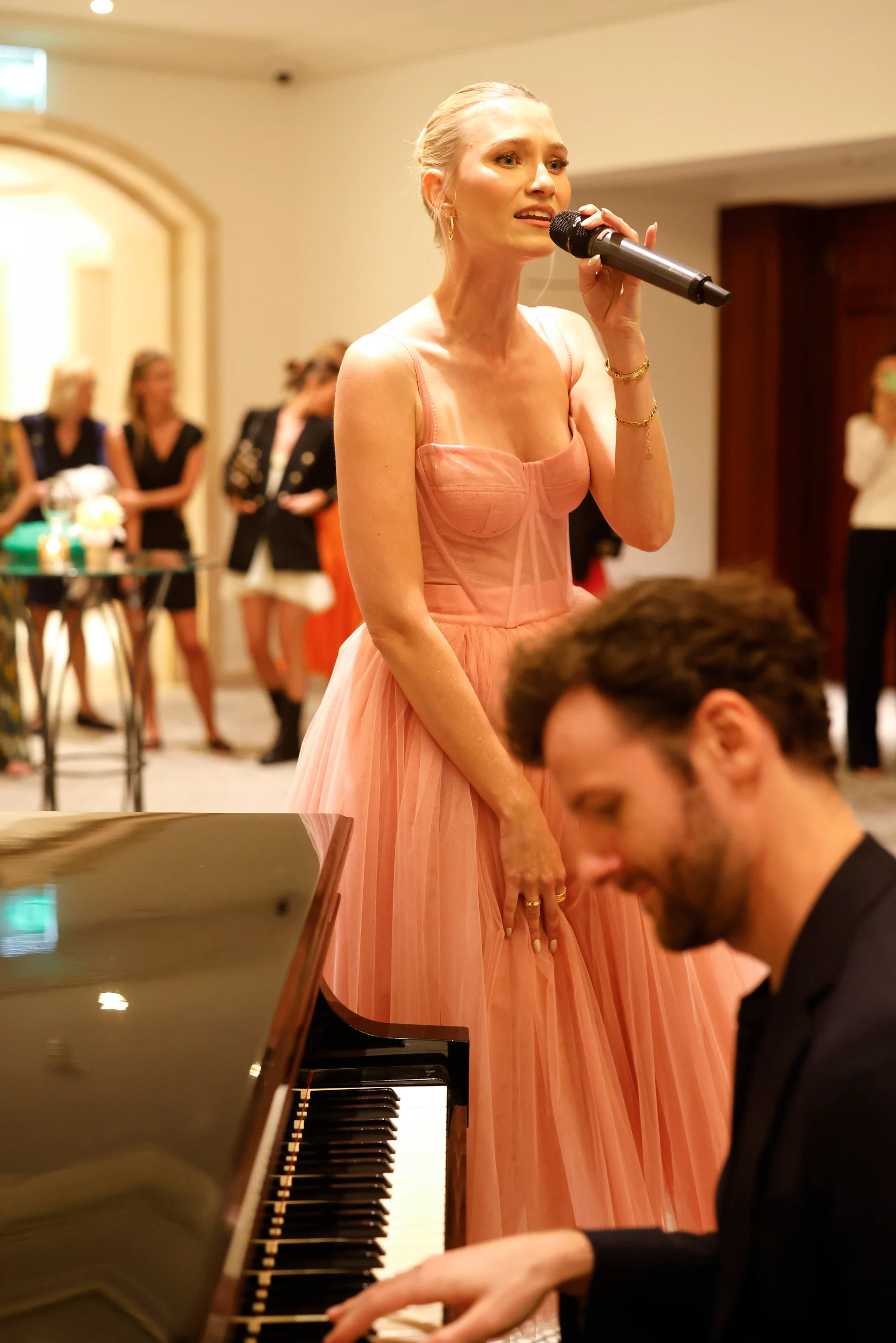 Sängerin im rosa Kleid singt bei einem Event neben einem Klavier, im Vordergrund sitzt ein Pianist.