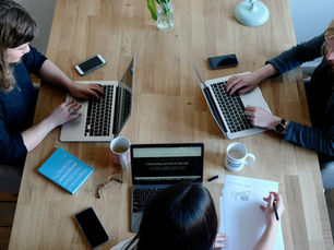 Drei Frauen sitzen an einem Holztisch, arbeiten an Laptops und Notizen, umgeben von Smartphones, Kaffee und Unterlagen.