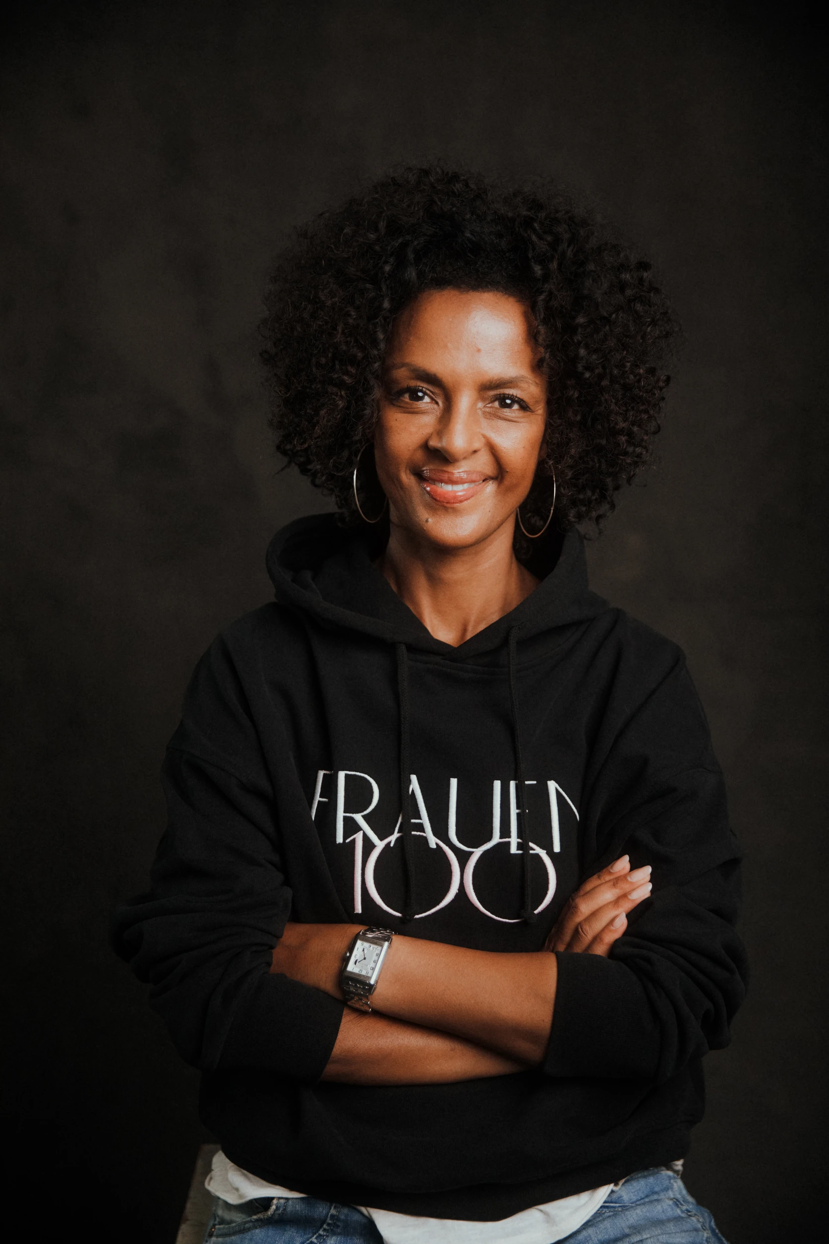 Frau mit lockigem Haar trägt einen schwarzen „Frauen100“-Hoodie und lächelt mit verschränkten Armen in die Kamera.