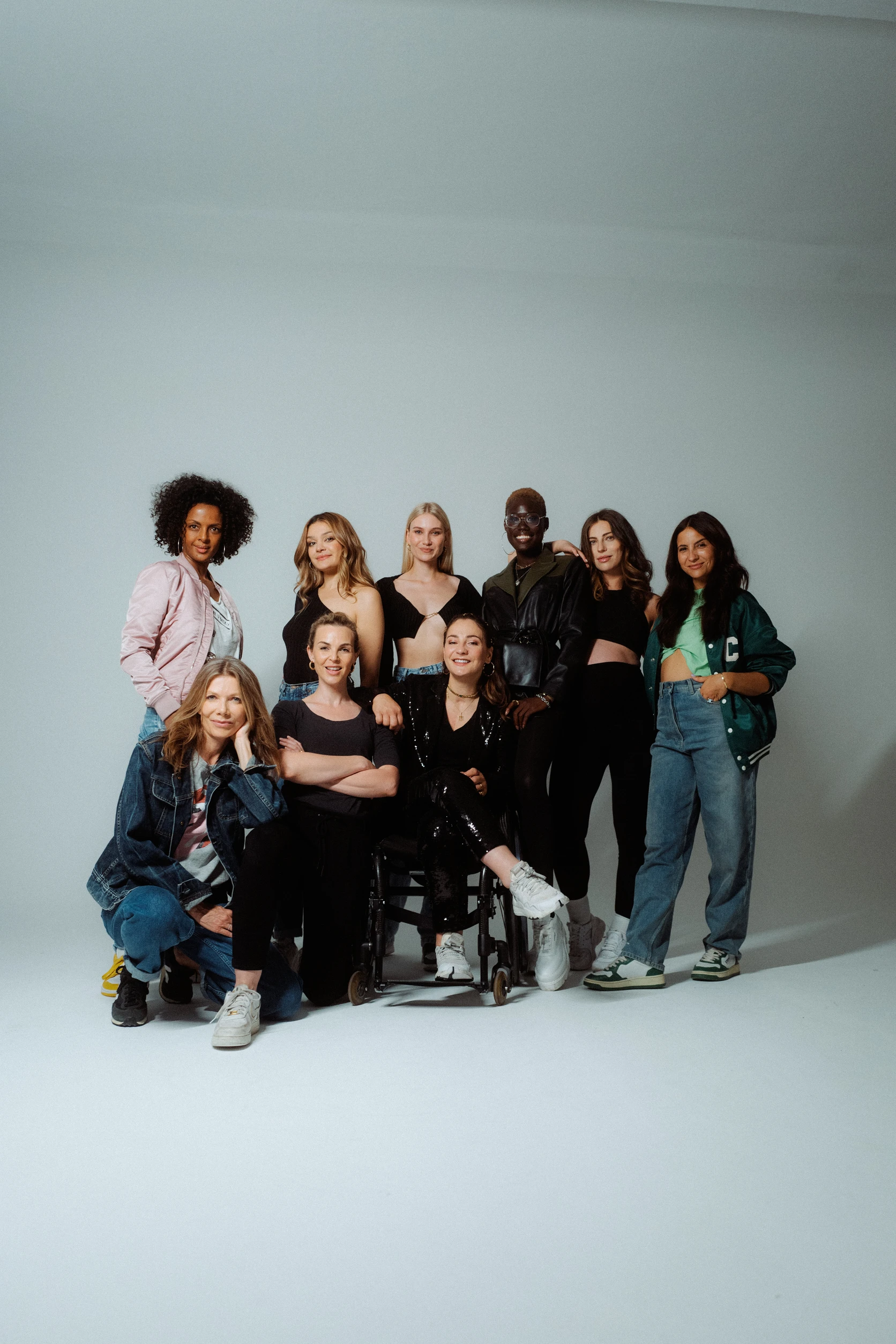 Diverse Gruppe von Frauen posiert gemeinsam im Studio vor hellem Hintergrund, eine Frau sitzt im Rollstuhl im Mittelpunkt.