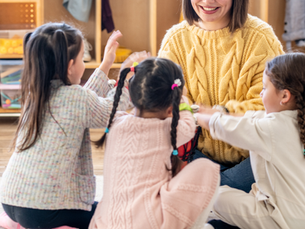Erzieherin sitzt mit drei kleinen Kindern im Kreis und spielt gemeinsam mit ihnen in einem Gruppenraum.