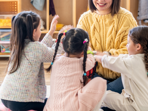Erzieherin sitzt mit drei kleinen Kindern im Kreis und spielt gemeinsam mit ihnen in einem Gruppenraum.