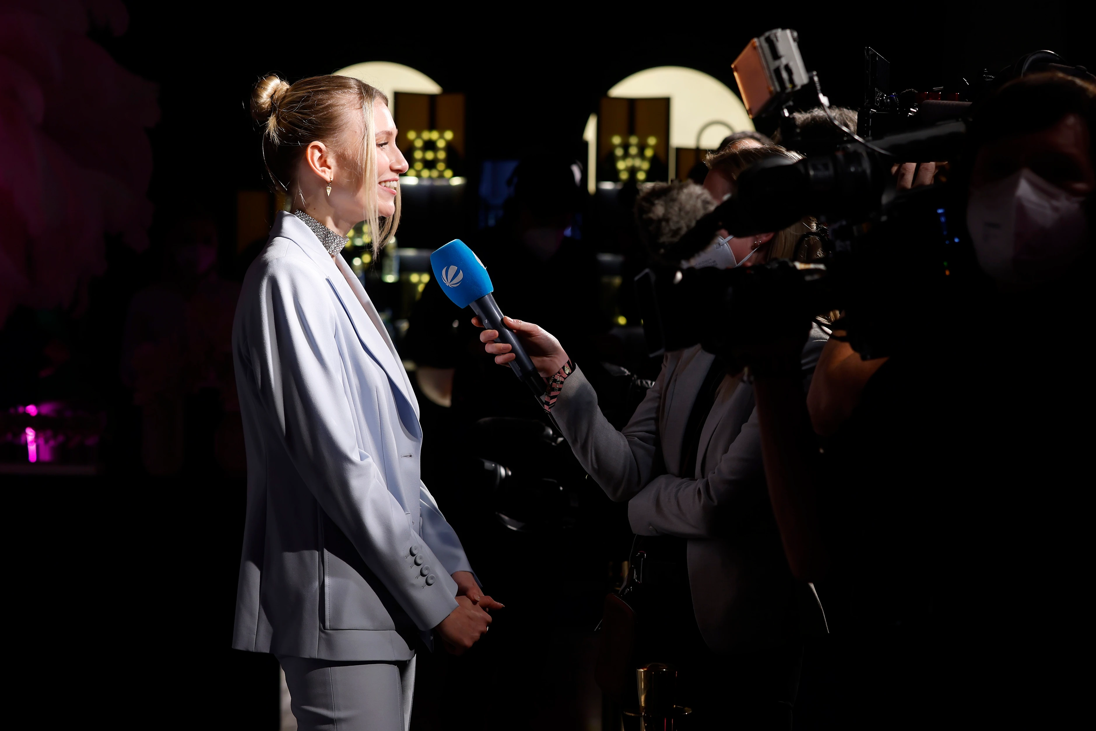 Frau im hellen Hosenanzug gibt ein Interview vor Kameras und Mikrofon auf einem Event.