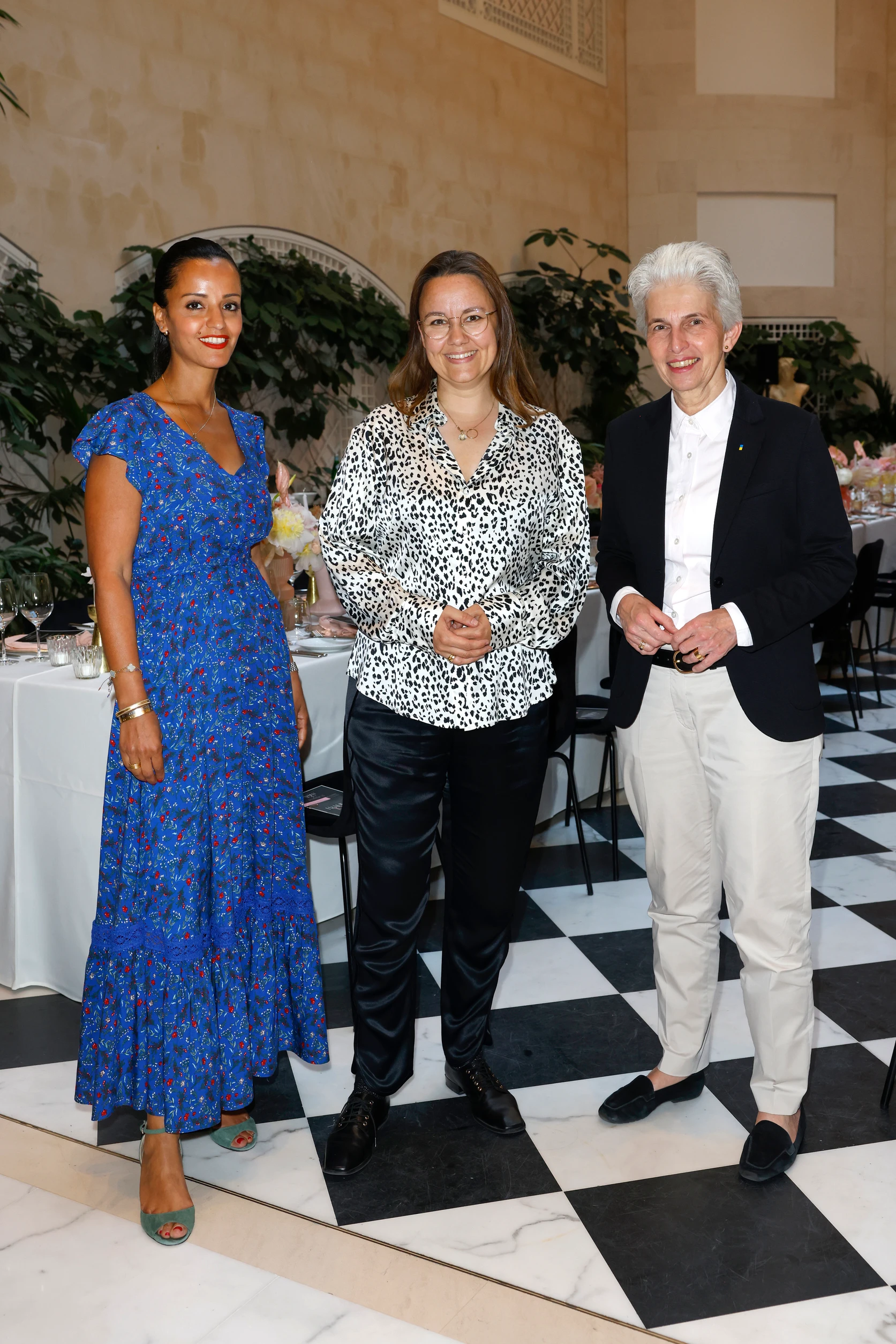 Drei Frauen stehen lächelnd in einem festlich dekorierten Raum mit gedeckten Tischen und Pflanzen im Hintergrund.