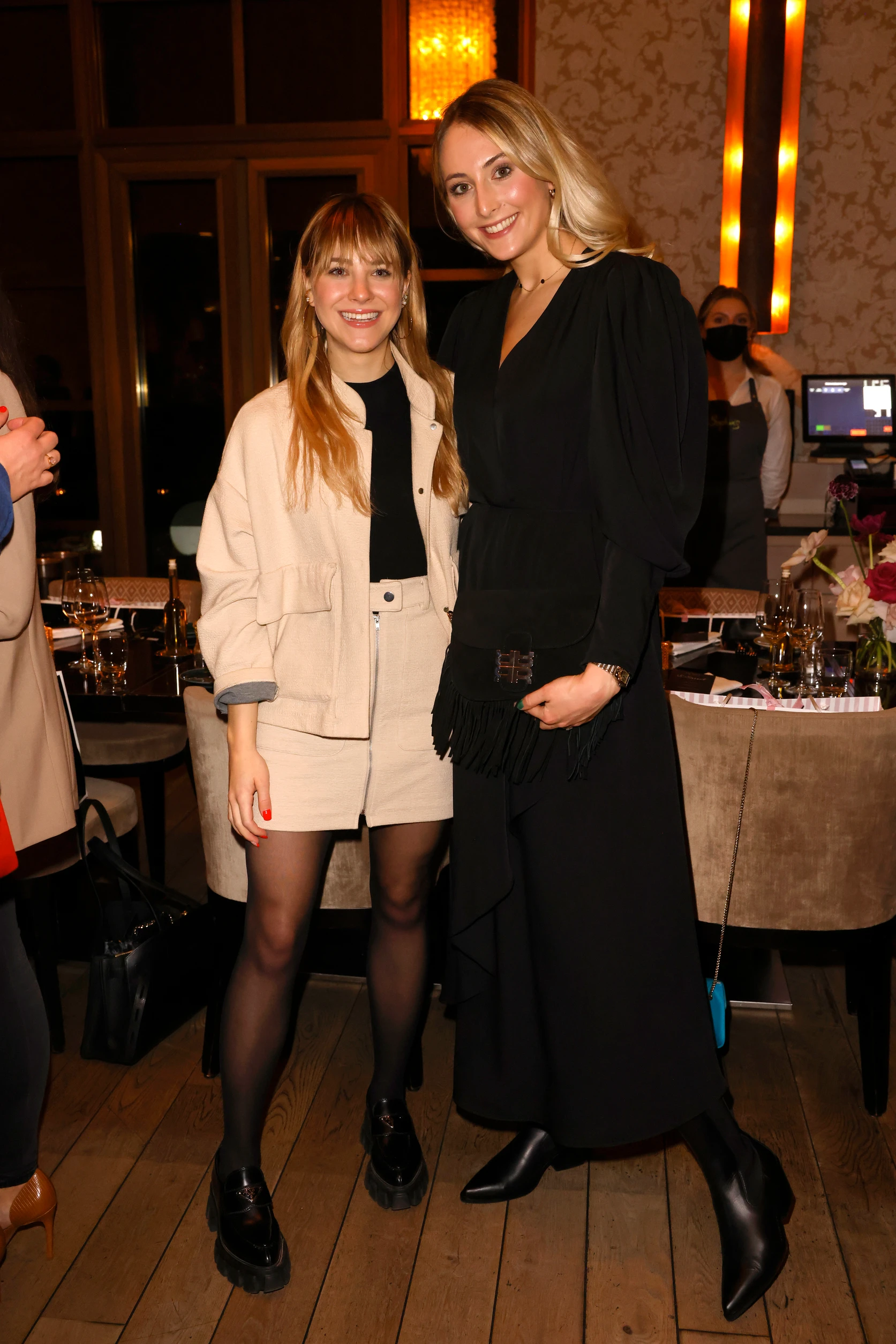 Zwei Frauen stehen bei einem Event nebeneinander und lächeln in die Kamera, elegant gekleidet vor einem Restaurant-Setting.