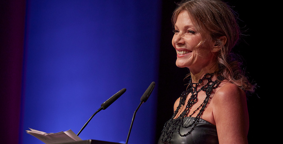 Eine Frau steht an einem Rednerpult, lächelt und hält eine Rede vor Publikum auf einer Bühne mit „FRAUEN100“-Beschriftung.