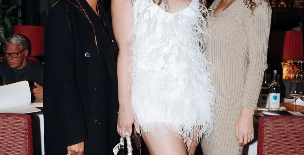 Drei Frauen stehen nebeneinander bei einem Event, eine trägt ein weißes Federkleid, die anderen dunkle und beige Outfits.