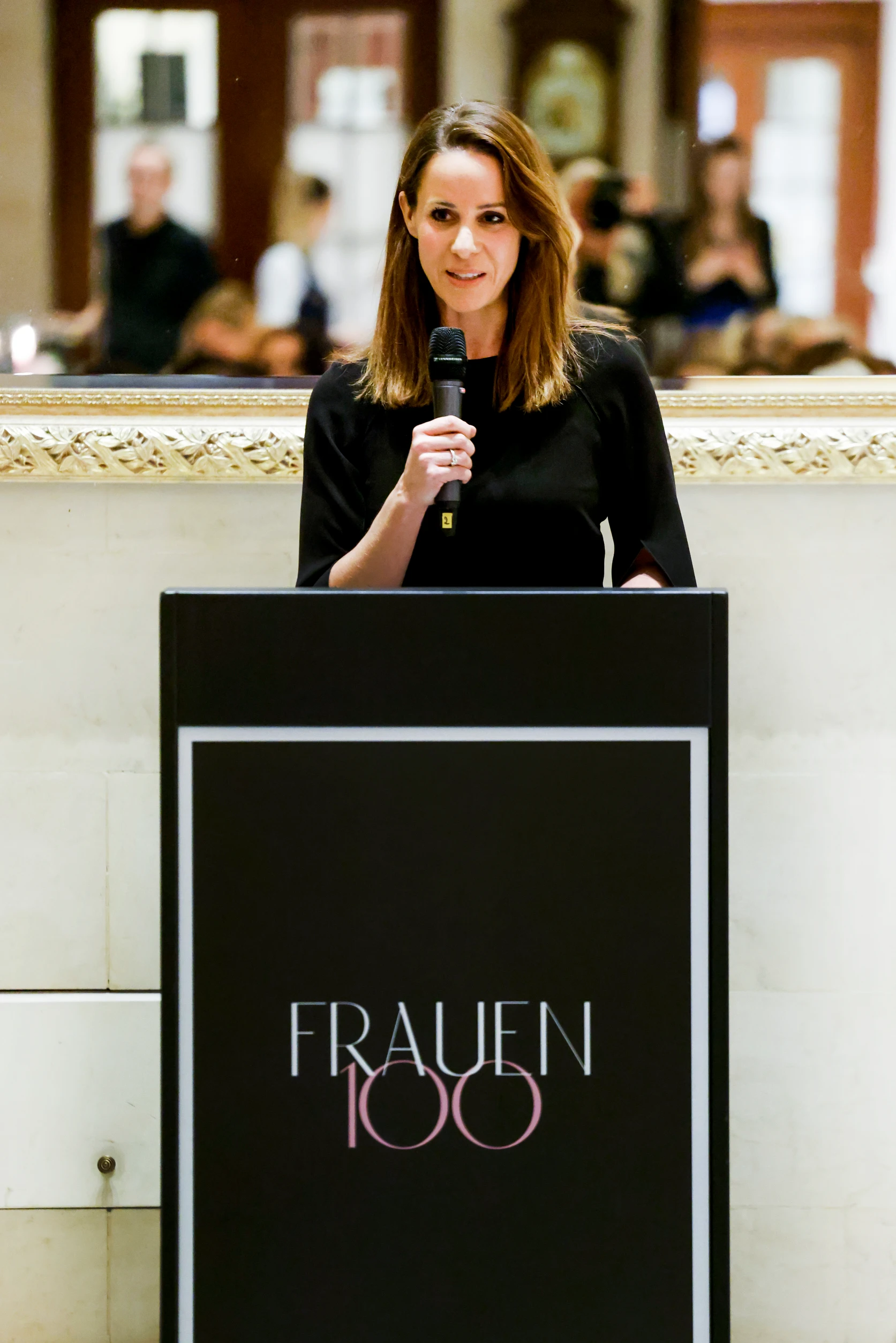 Frau steht an einem Rednerpult mit der Aufschrift „FRAUEN 100“ und spricht mit einem Mikrofon vor Publikum.
