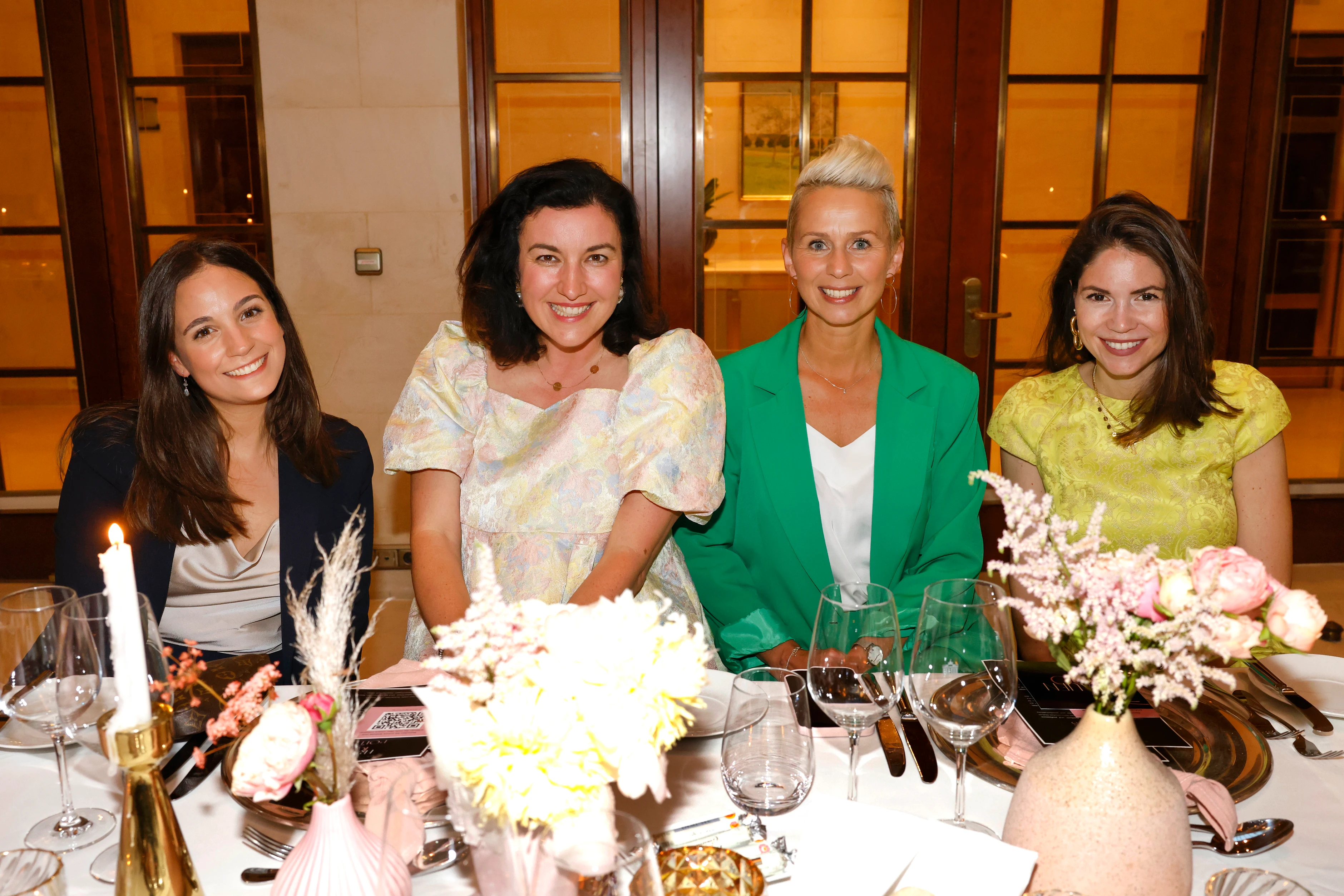 Vier Frauen sitzen gemeinsam an einem festlich gedeckten Tisch mit Blumen und Kerzen und lächeln in die Kamera.