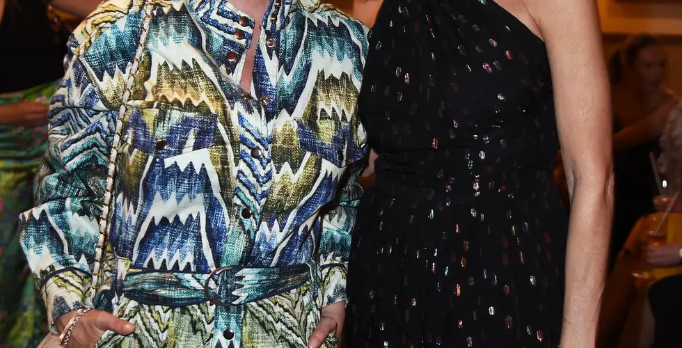 Zwei Frauen stehen nebeneinander bei einem Event und lächeln in die Kamera; eine trägt einen gemusterten Jumpsuit, die andere ein schwarzes, schulterfreies Outfit, im Hintergrund ein stilvoll beleuchteter Raum.