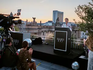 Frau steht am Rednerpult und gegenüber befindet sich die Presse. Das Ganze bei untergehender Sonne. 