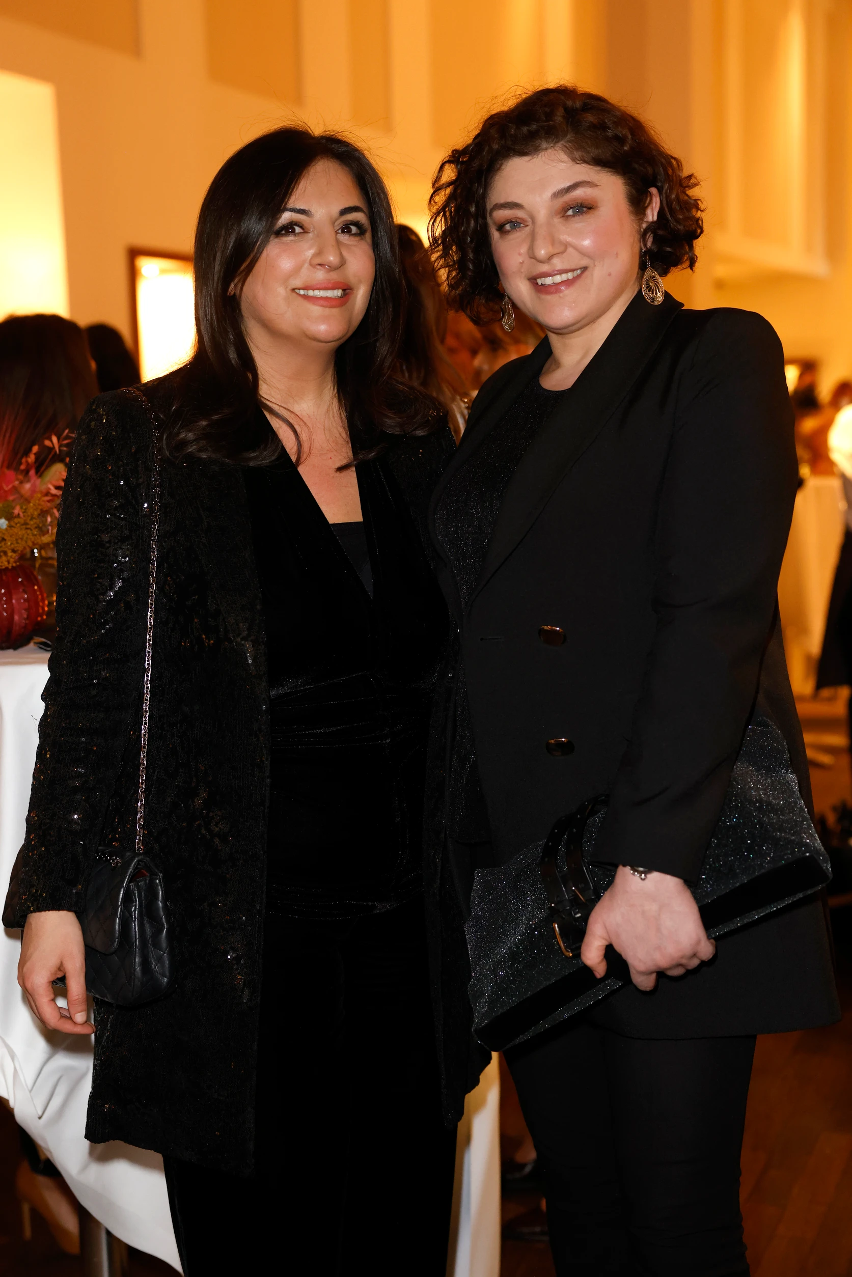 Zwei Frauen stehen nebeneinander bei einem Event, lächeln in die Kamera und tragen elegante schwarze Outfits, im Hintergrund warm beleuchteter Innenraum mit Gästen.
