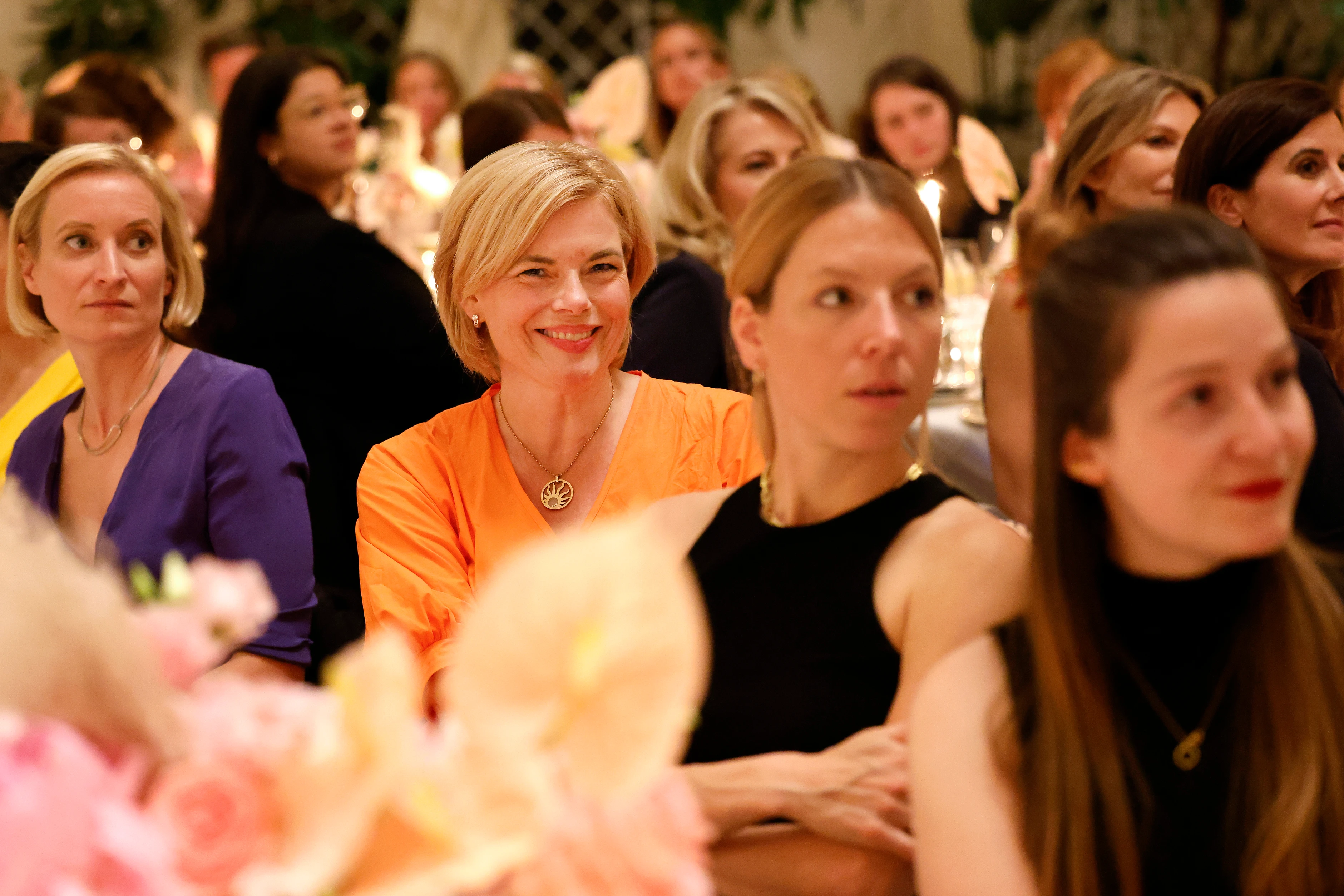 Mehrere Frauen sitzen an gedeckten Tischen, im Fokus eine Frau im orangenen Kleid, die lächelt.