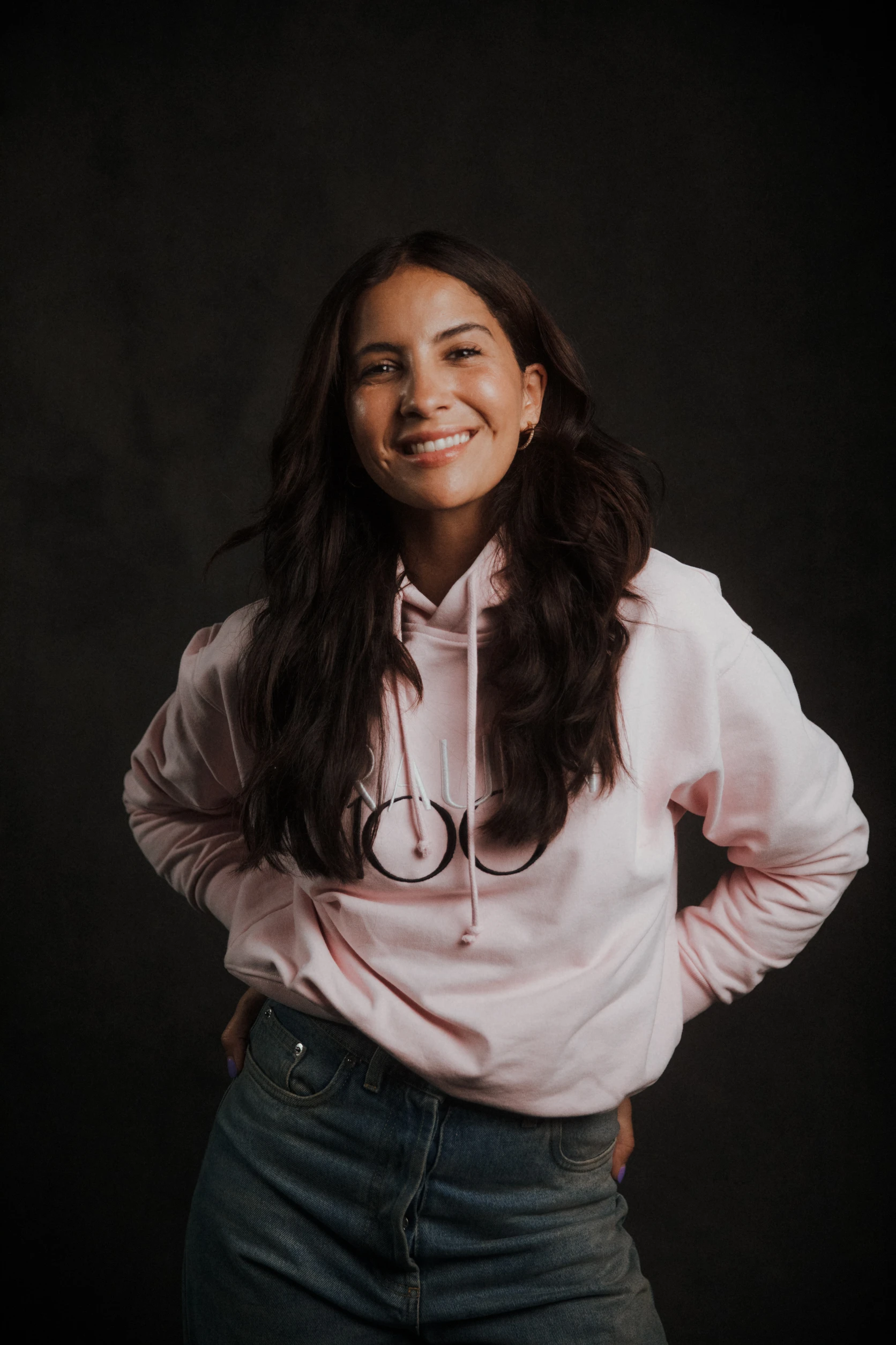 Frau im rosa Hoodie lächelt in die Kamera und steht locker mit Händen in der Hüfte vor dunklem Hintergrund.