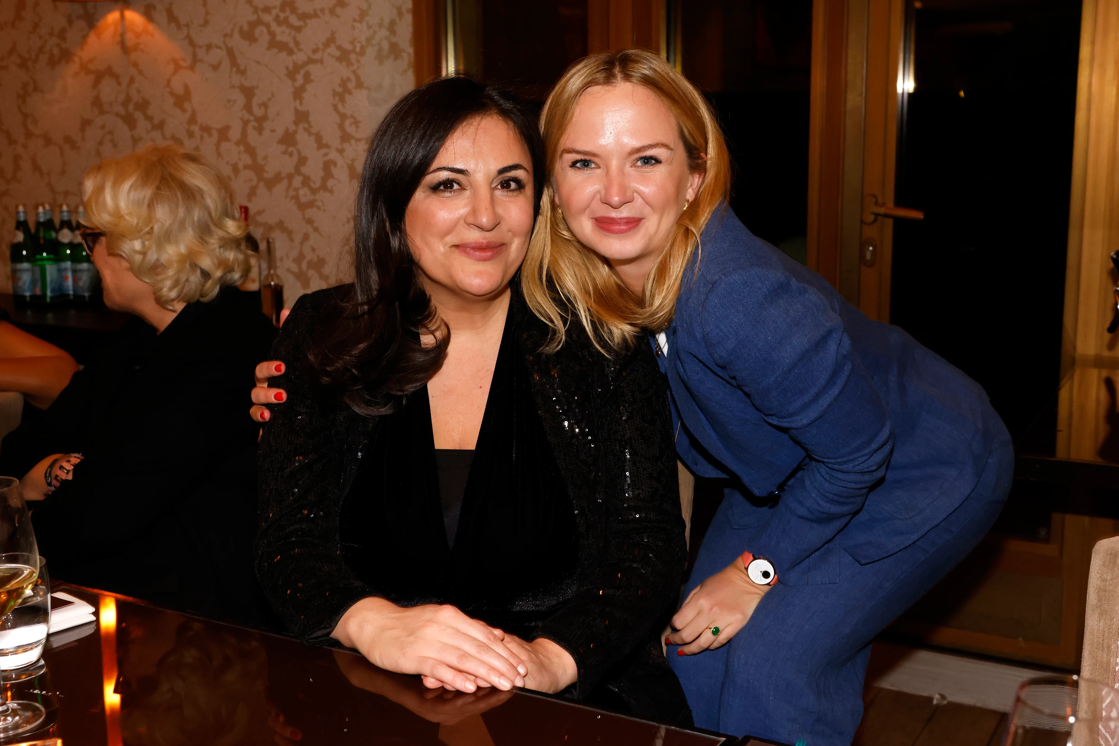 Zwei Frauen sitzen eng zusammen an einem Tisch und lächeln in die Kamera, eine legt freundschaftlich den Arm um die andere.