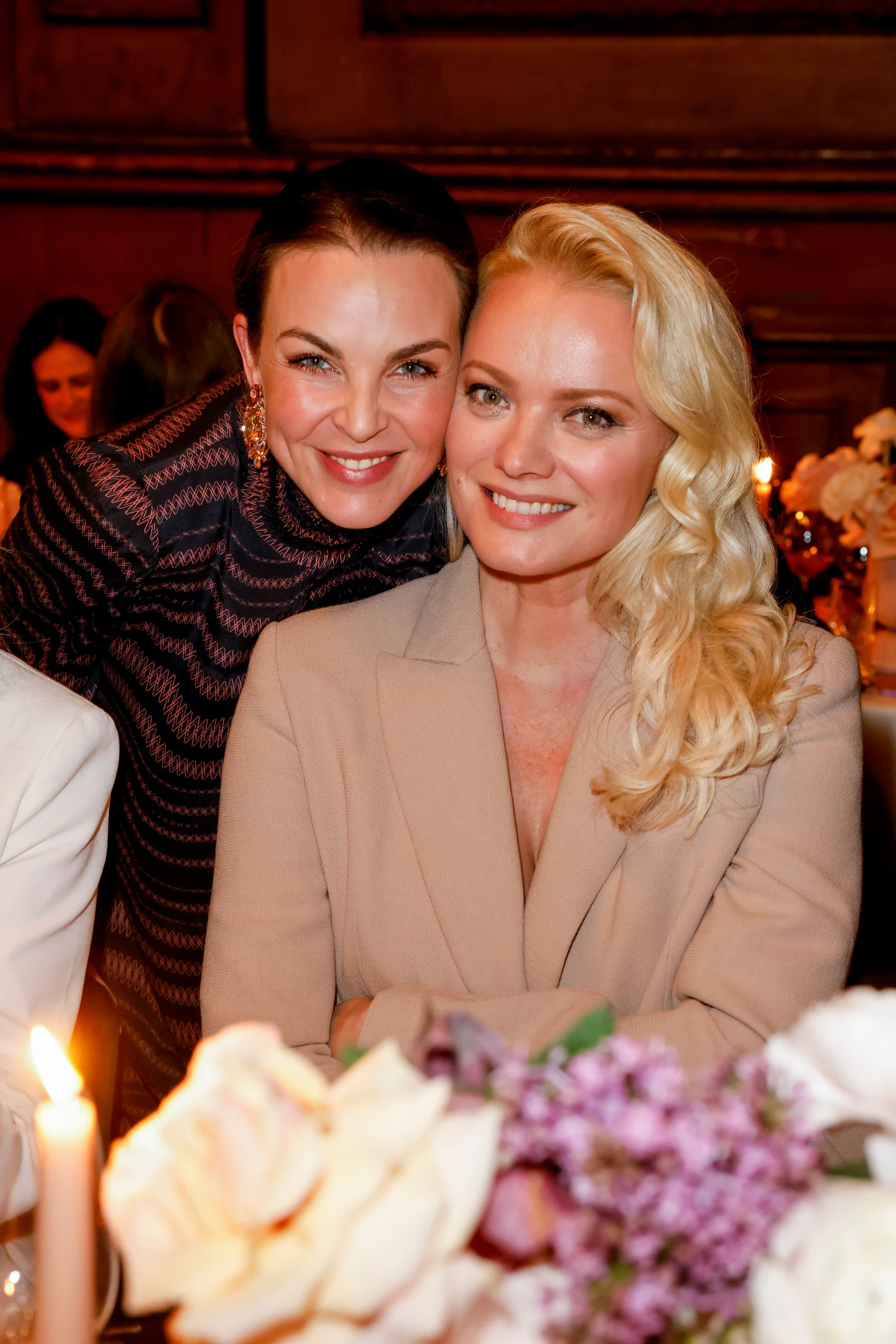 Zwei Frauen sitzen eng zusammen bei einem Dinner-Event und lächeln in die Kamera.
