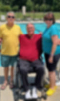 Three adults smile by a sunny poolside, one seated in a wheelchair—capturing the spirit of community and inclusion behind our swimming fundraiser for seniors.