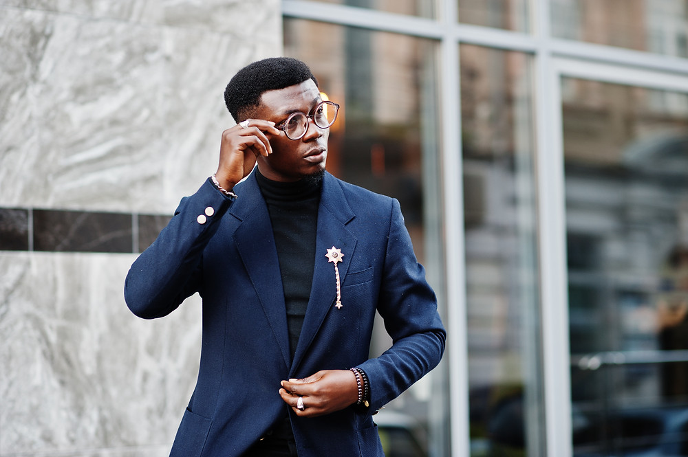 A dapper black man dressed in a navy blue suit with black tshirt and gold accessories