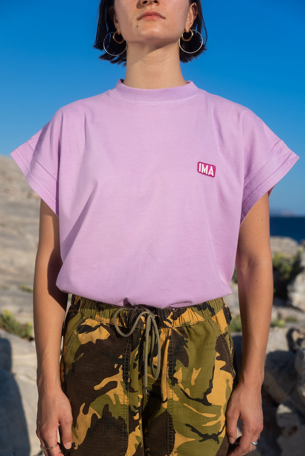 Thumbnail: Irregular Movement of Arts: Fay Tzouma wearing the I.M.A Fenily Orchid t-shirt, in lilac, which features an illustration on the back, designed by artist Aristomenis Theodoropoulos.