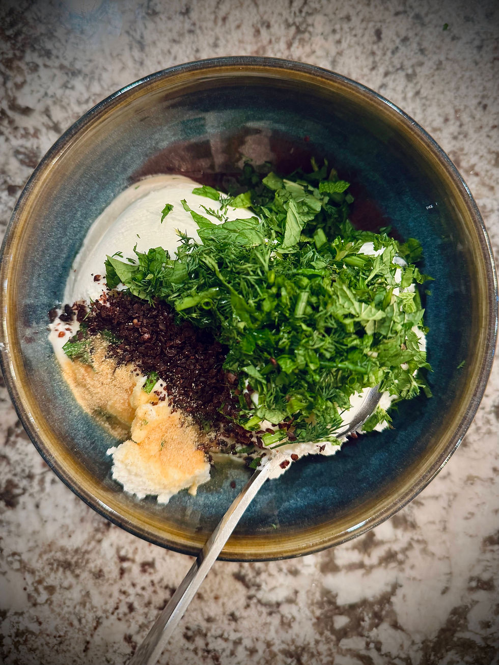 A bowl with yogurt, chopped herbs, and spices on a marbled counter. A spoon is inside the bowl. The setting is rustic and earthy.