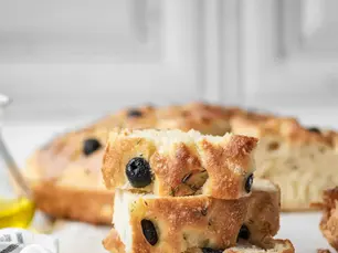 Freshly baked gluten-free focaccia made with Fioreglut flour, sliced and stacked on parchment paper with black olives and rosemary, next to a drizzle of olive oil and a white linen towel.